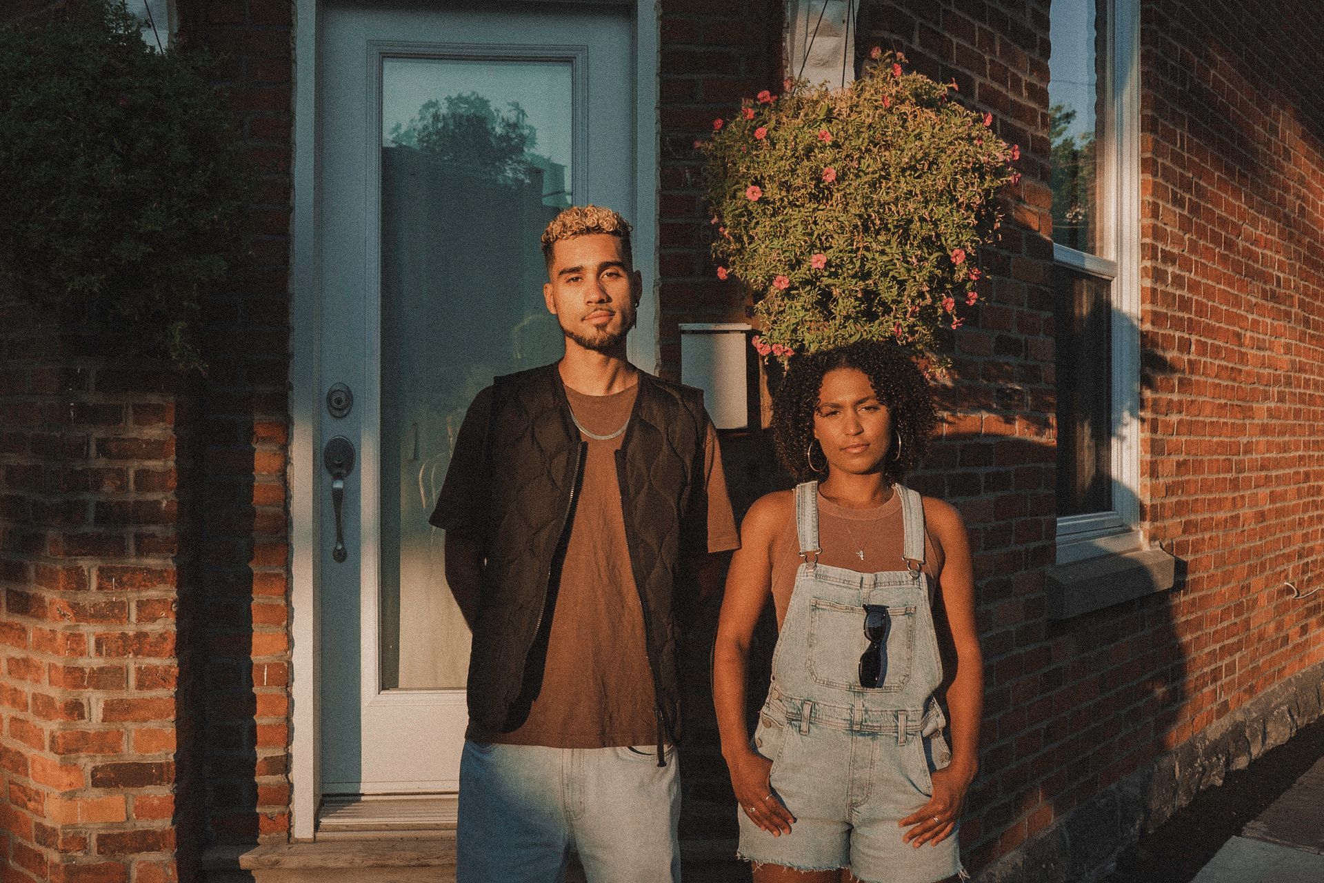 Two people stand outside a brick building. One wears a brown shirt and black vest, the other overalls. Sunny day.