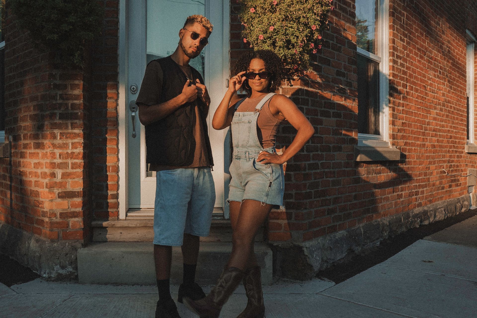 Two people stand outside a brick building. One wears overalls and sunglasses, the other a jacket and shorts.