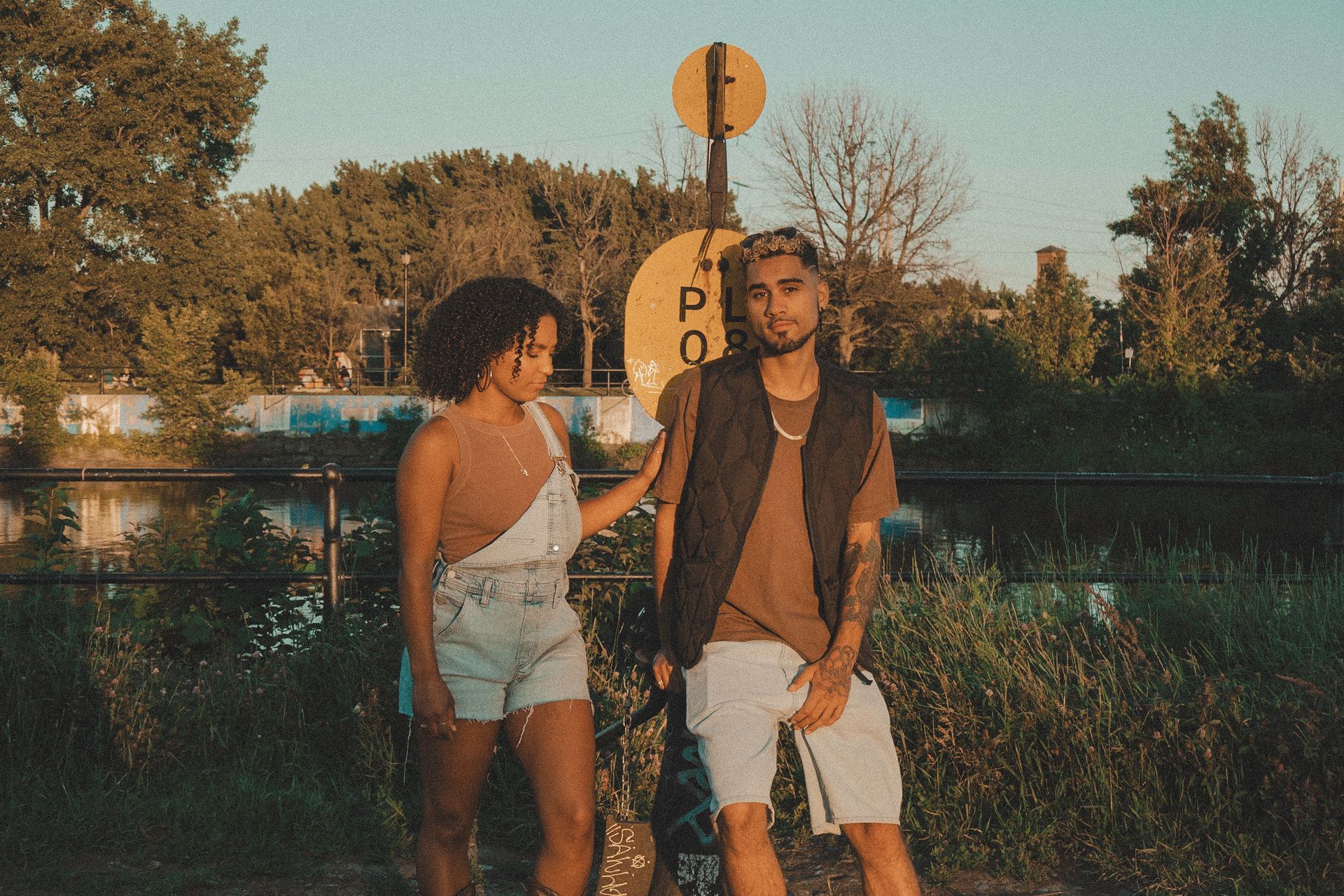 Two people by a yellow sign next to a body of water; woman in overalls, man in vest.