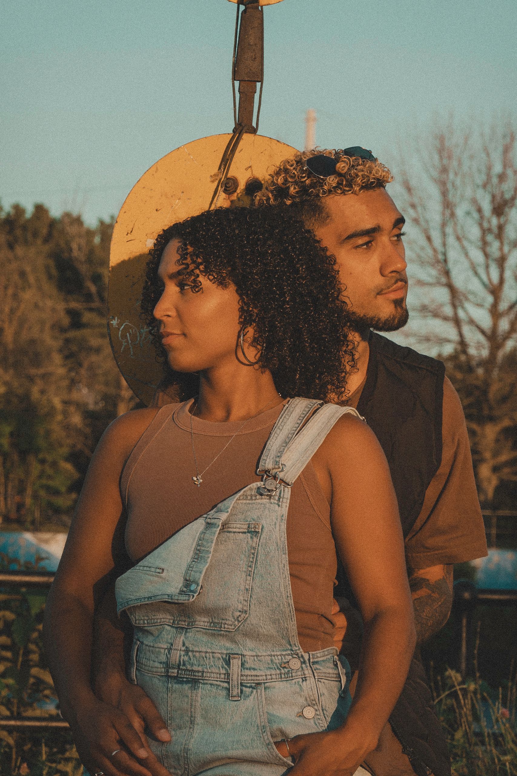 Couple embracing outdoors, woman in overalls, man behind her. Warm lighting, rustic setting.