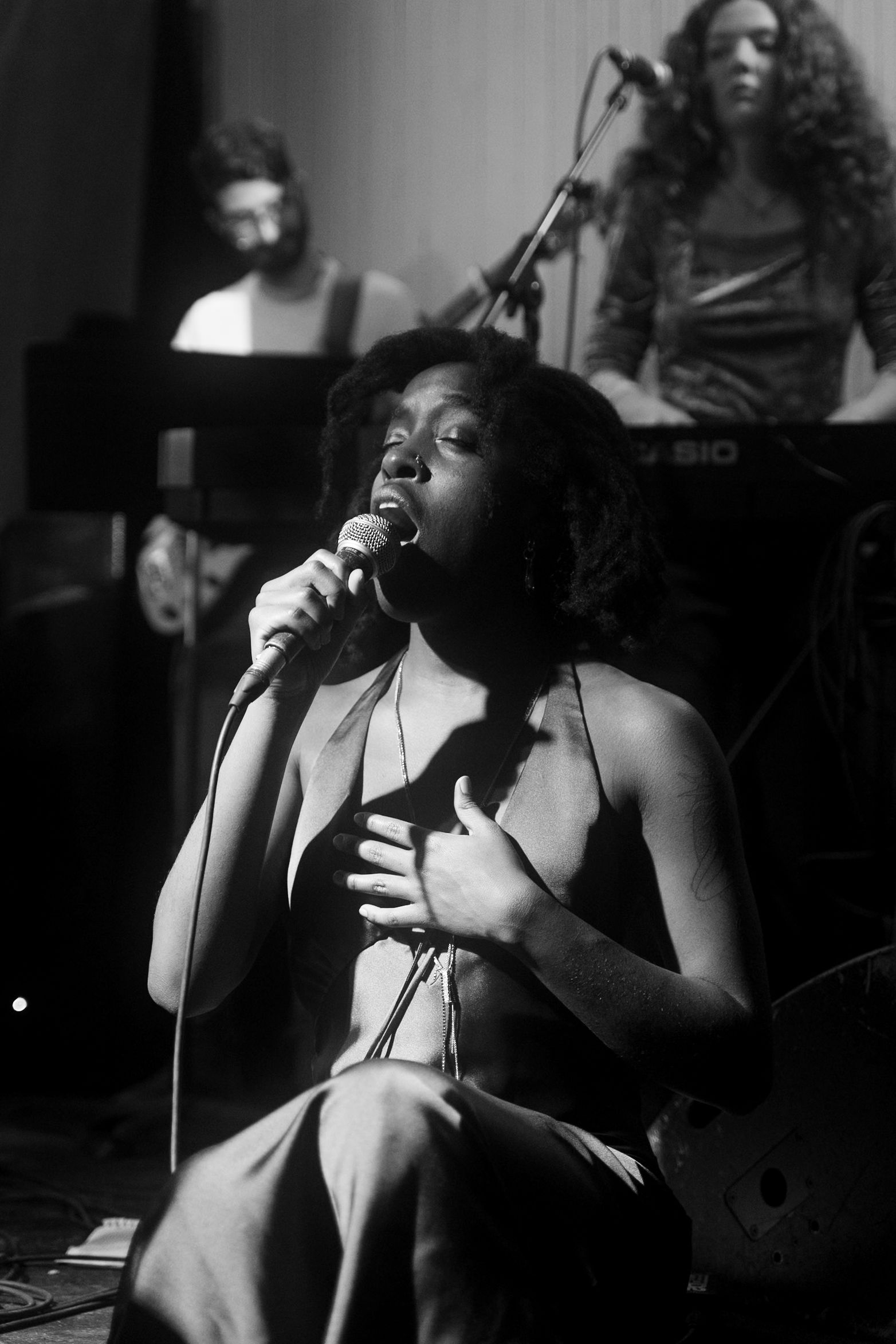 Singer performing on stage, holding microphone, hand on chest, with band members in background.