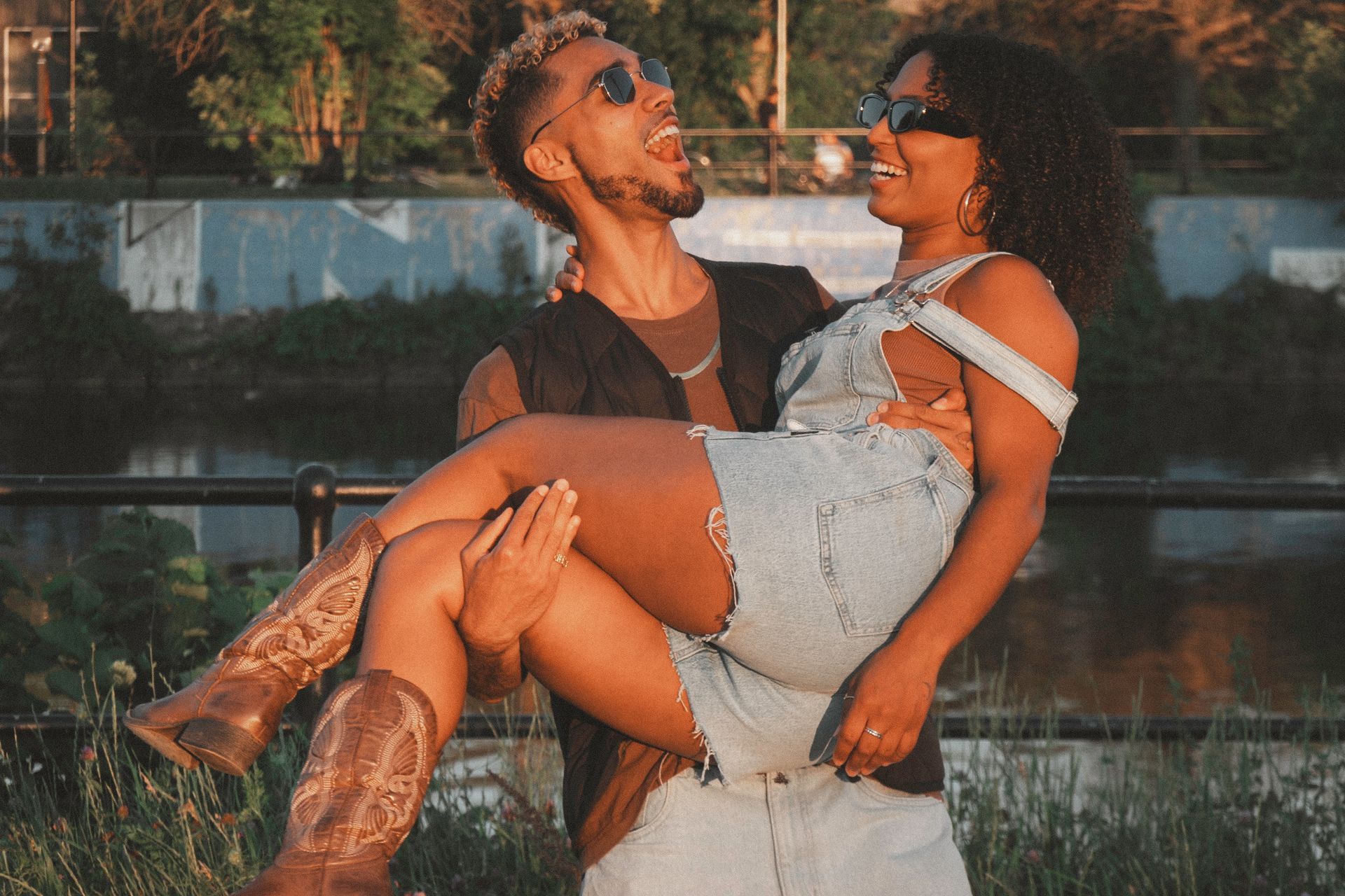Man carrying woman, both laughing and wearing sunglasses; outdoors, near water.