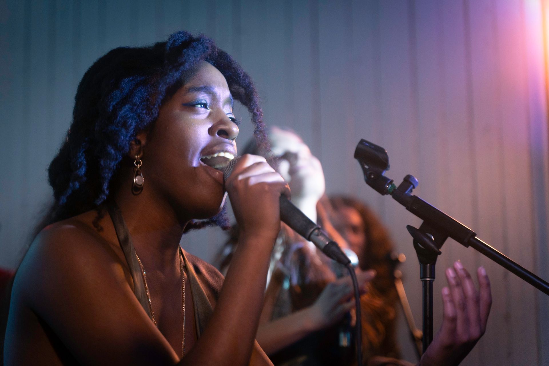 Woman singing into a microphone, lit by stage lights, two other singers blurred in the background.