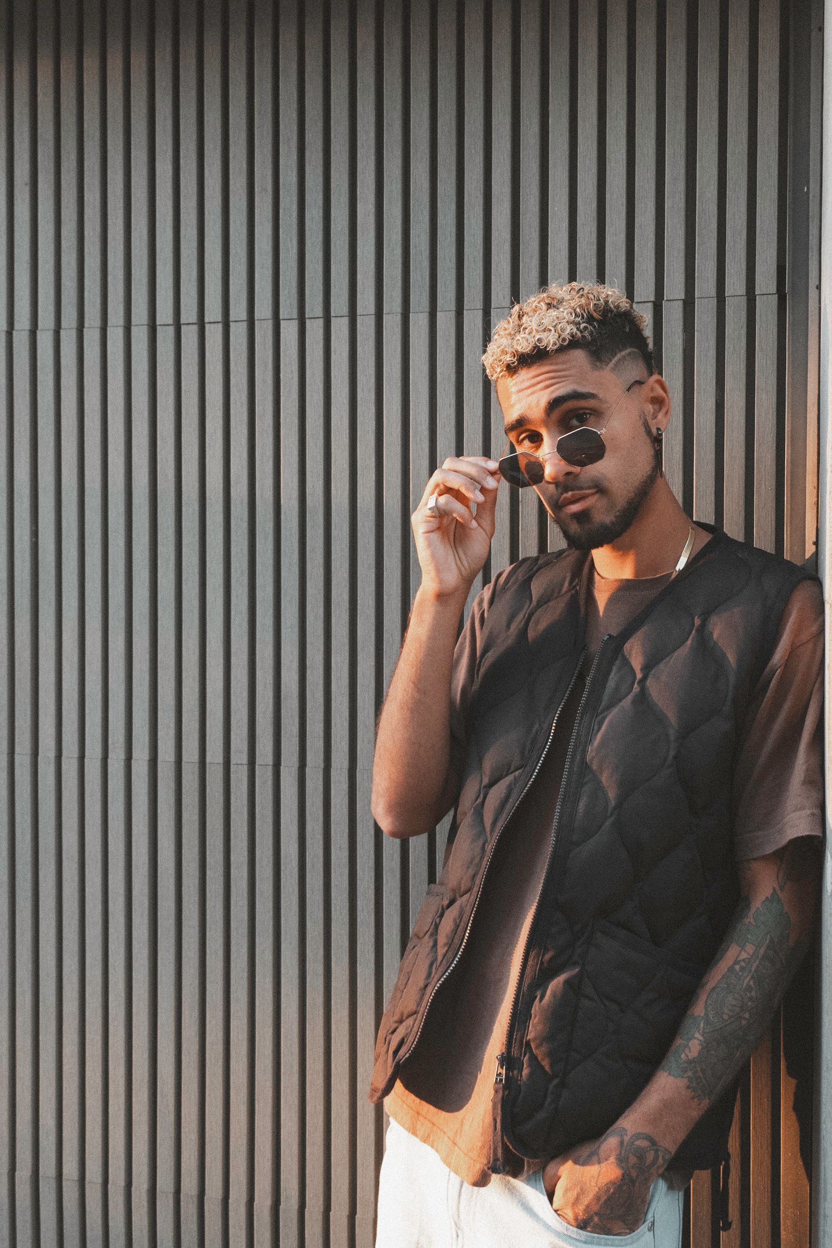 Man in vest and sunglasses leaning against a ribbed wall, adjusting glasses.