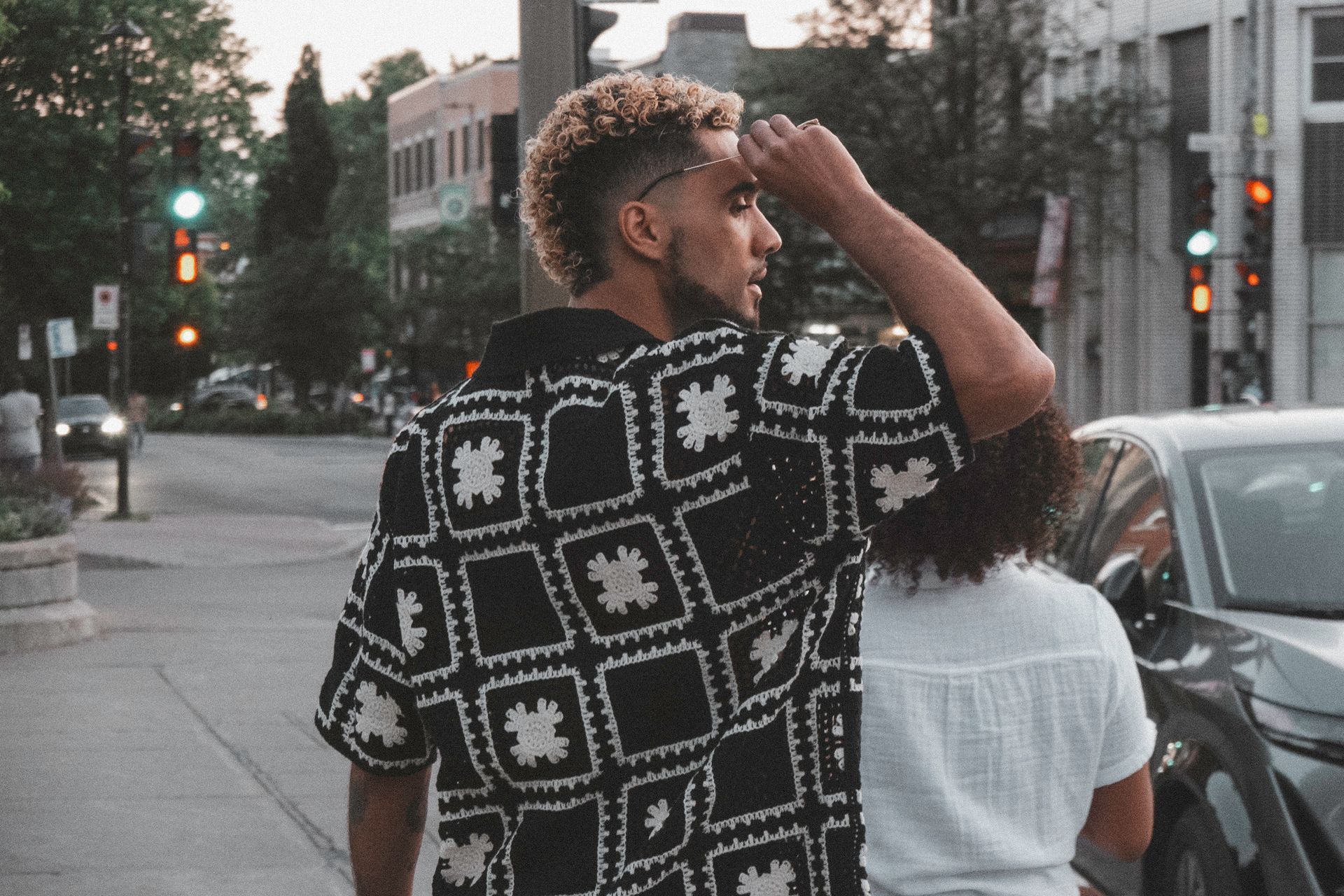 Man in patterned shirt on city street, looking away. Another person near car.