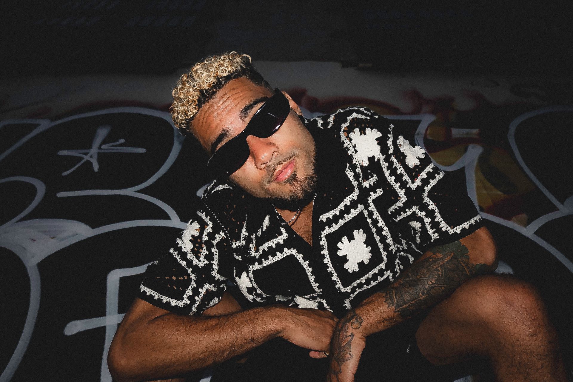Man wearing sunglasses and patterned shirt, squatting against a graffiti-covered wall.