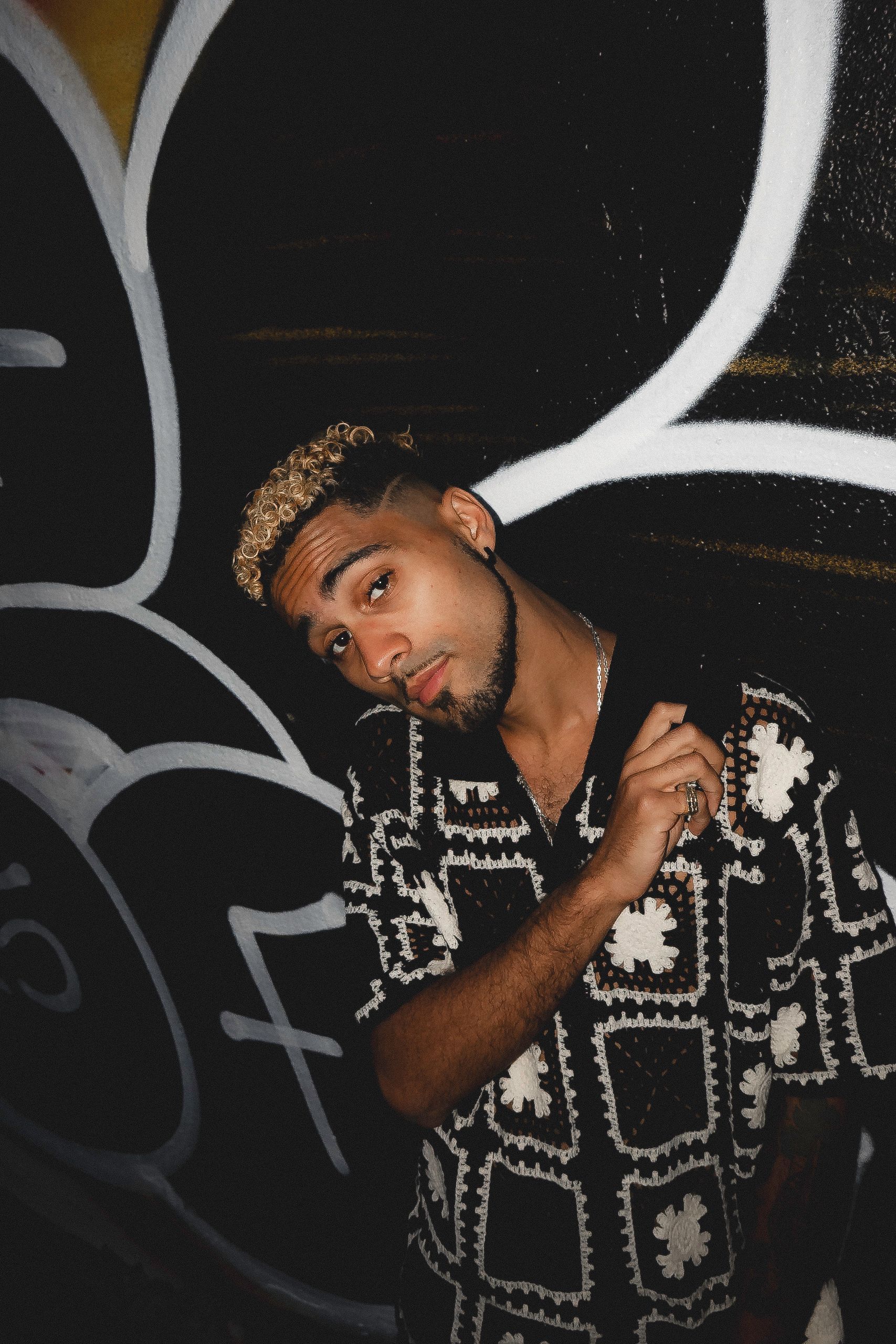 Man in patterned shirt leaning against graffiti, looking at the camera.