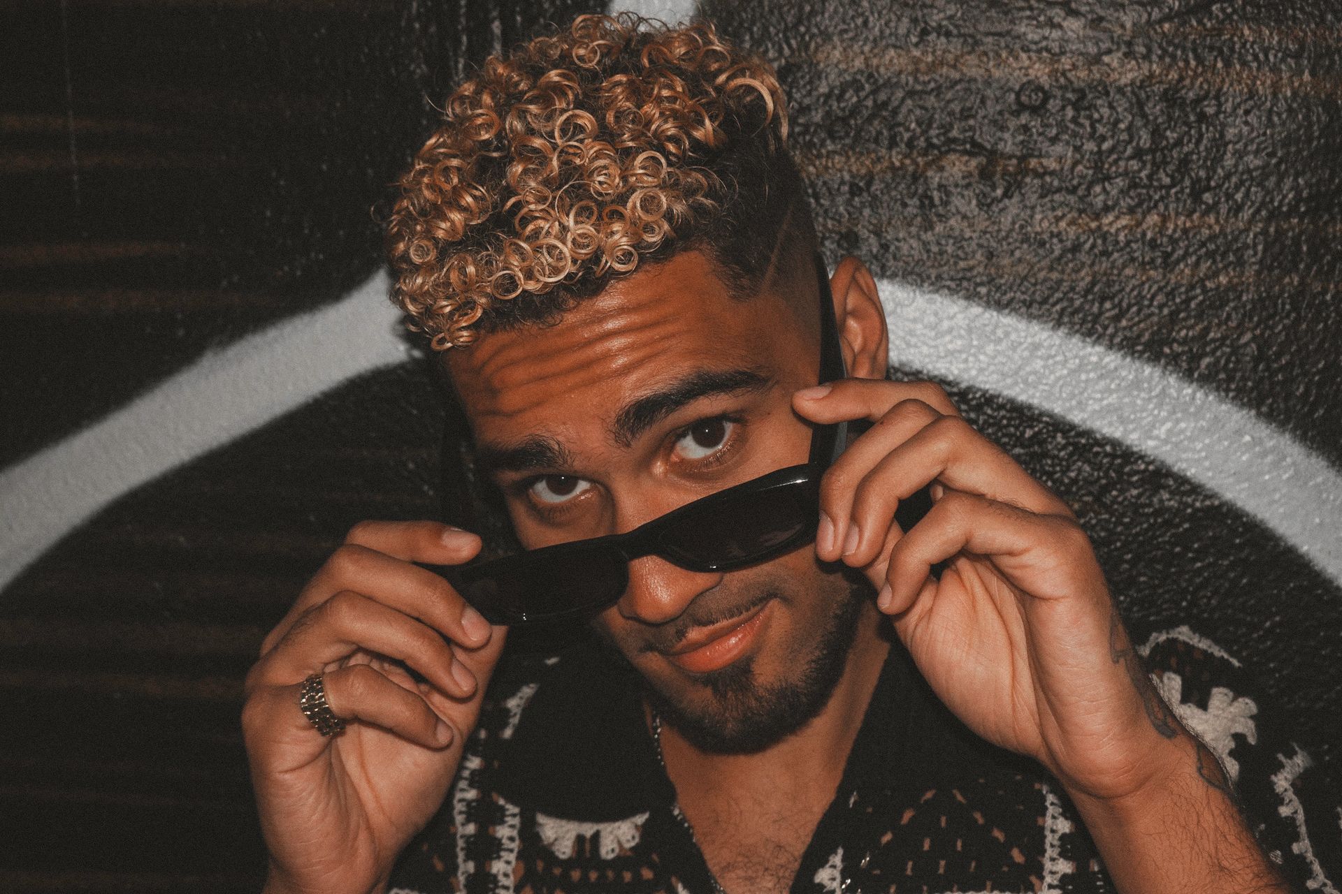 Man in sunglasses, holding them up slightly, with blonde curly hair. Leaning against a graffiti wall.