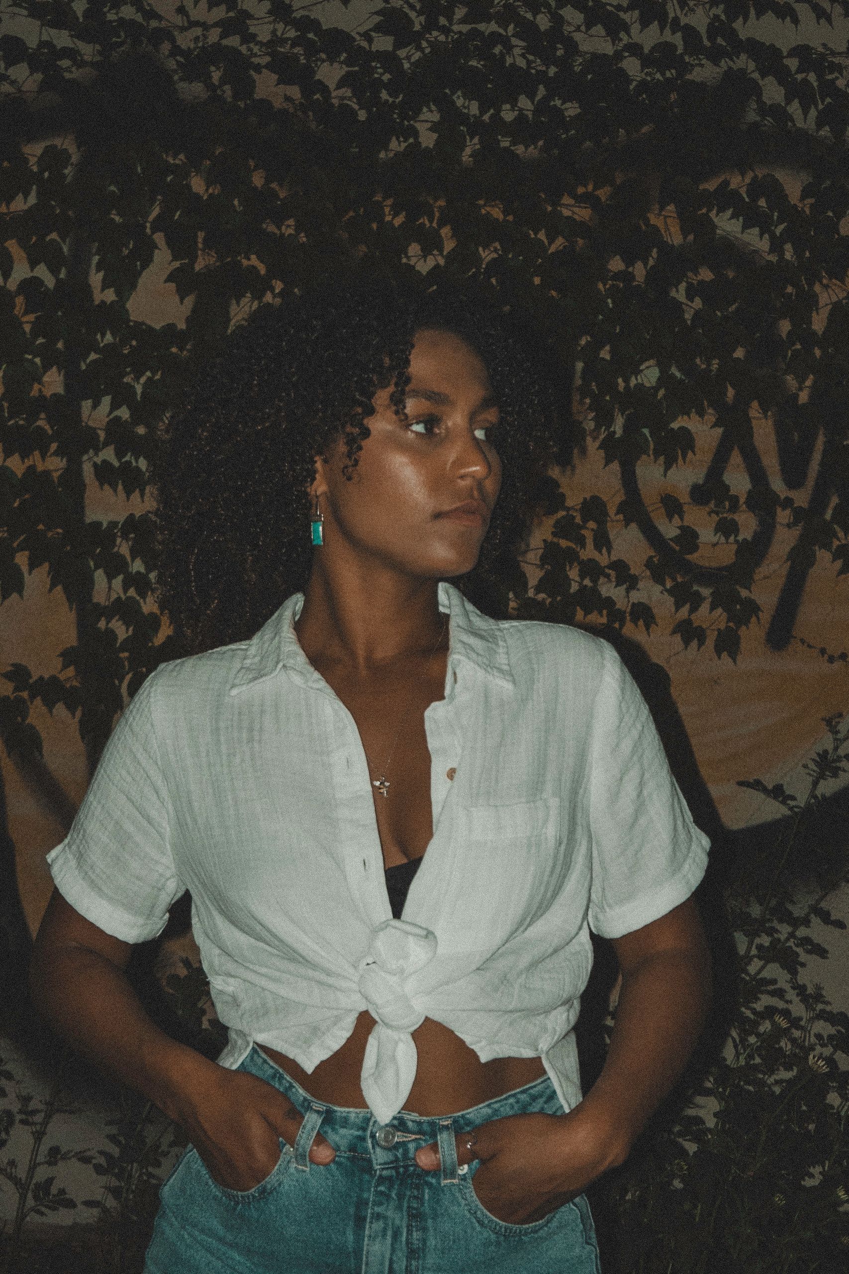 Woman in white tied shirt and jeans, hands in pockets, posing outdoors.