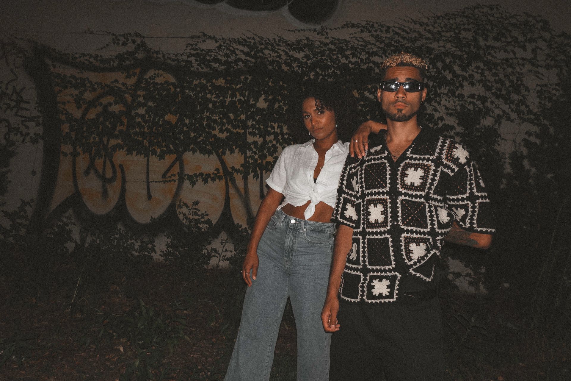 Woman in tied white shirt and jeans with man in patterned shirt and sunglasses, in front of graffiti.