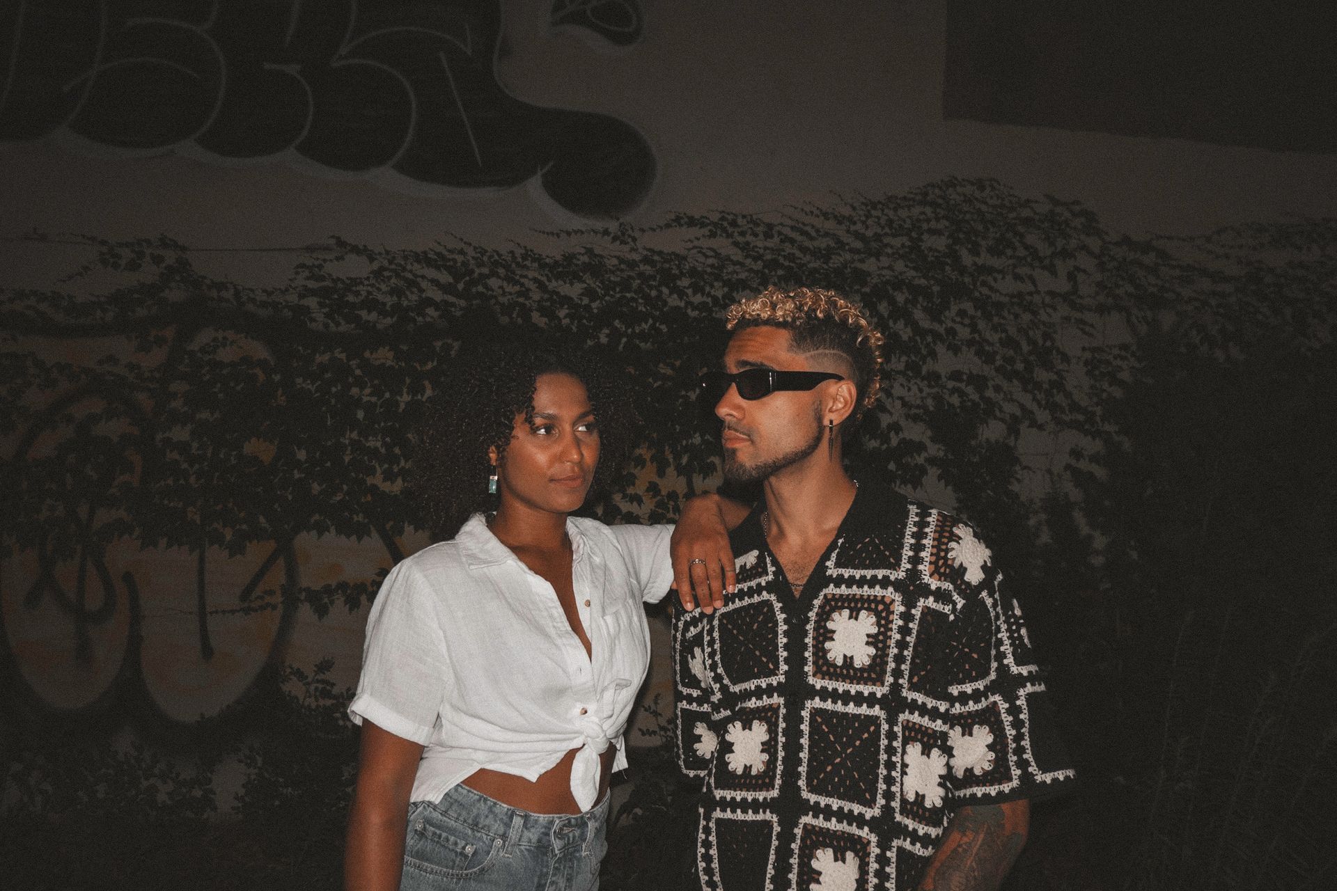 Woman in white shirt and man in patterned shirt pose in front of graffiti.