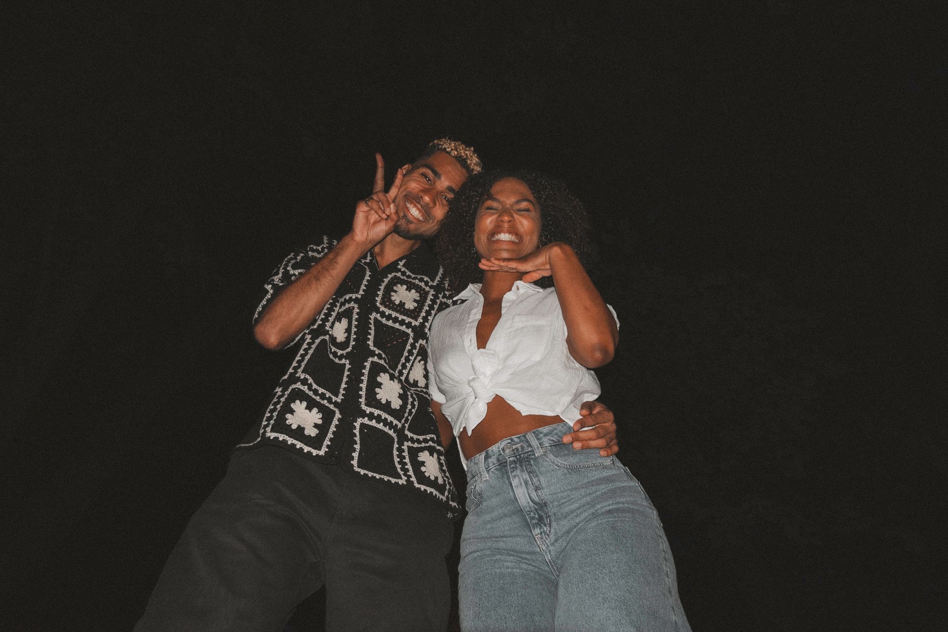 Two people smiling, posing in front of a dark background. The person on the left is wearing a patterned shirt. The person on the right has curly hair.