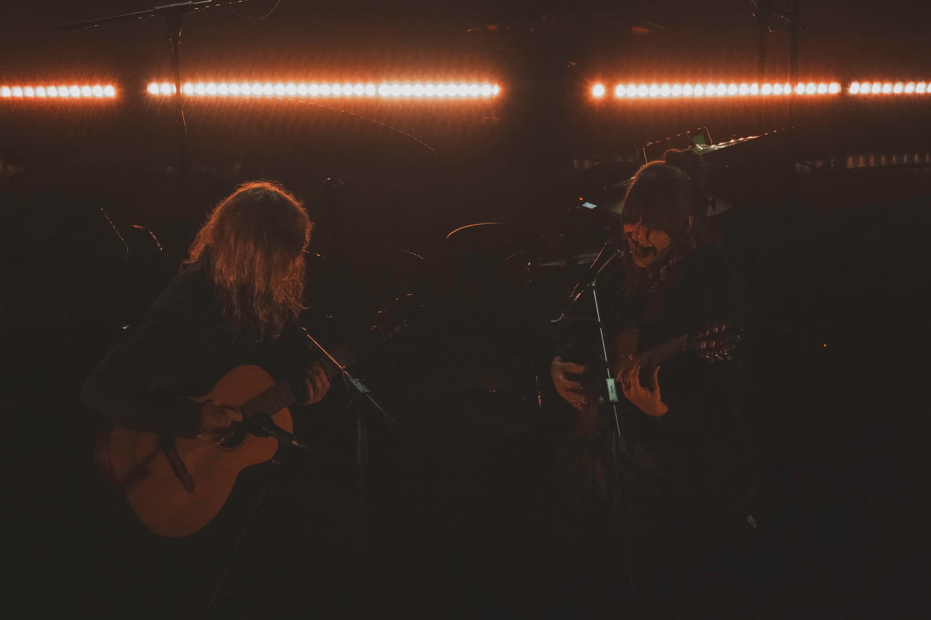 Two musicians performing on stage, lit by warm orange lights. One sings, plays acoustic guitar. The other plays electric guitar.