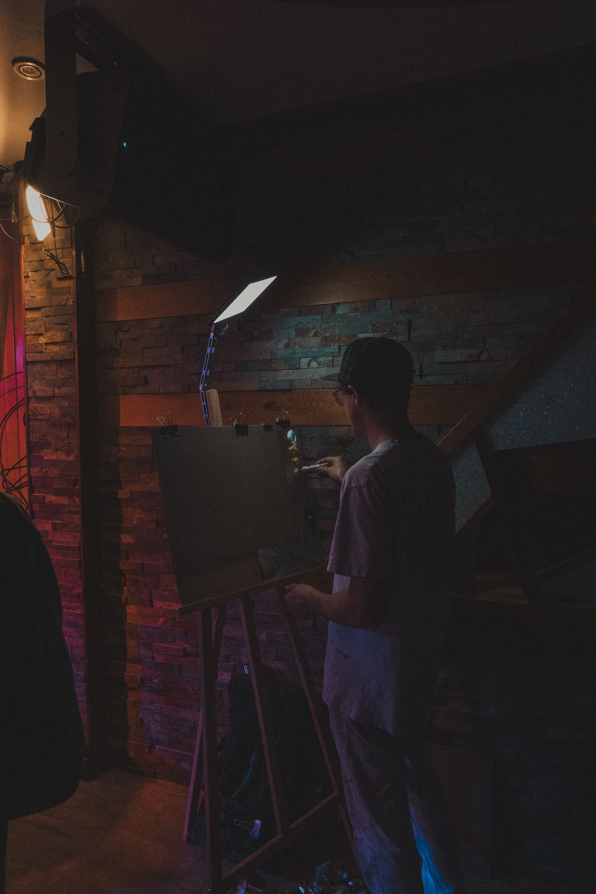 Person painting at an easel with bright lights, indoors, against a dark brick and wood background.