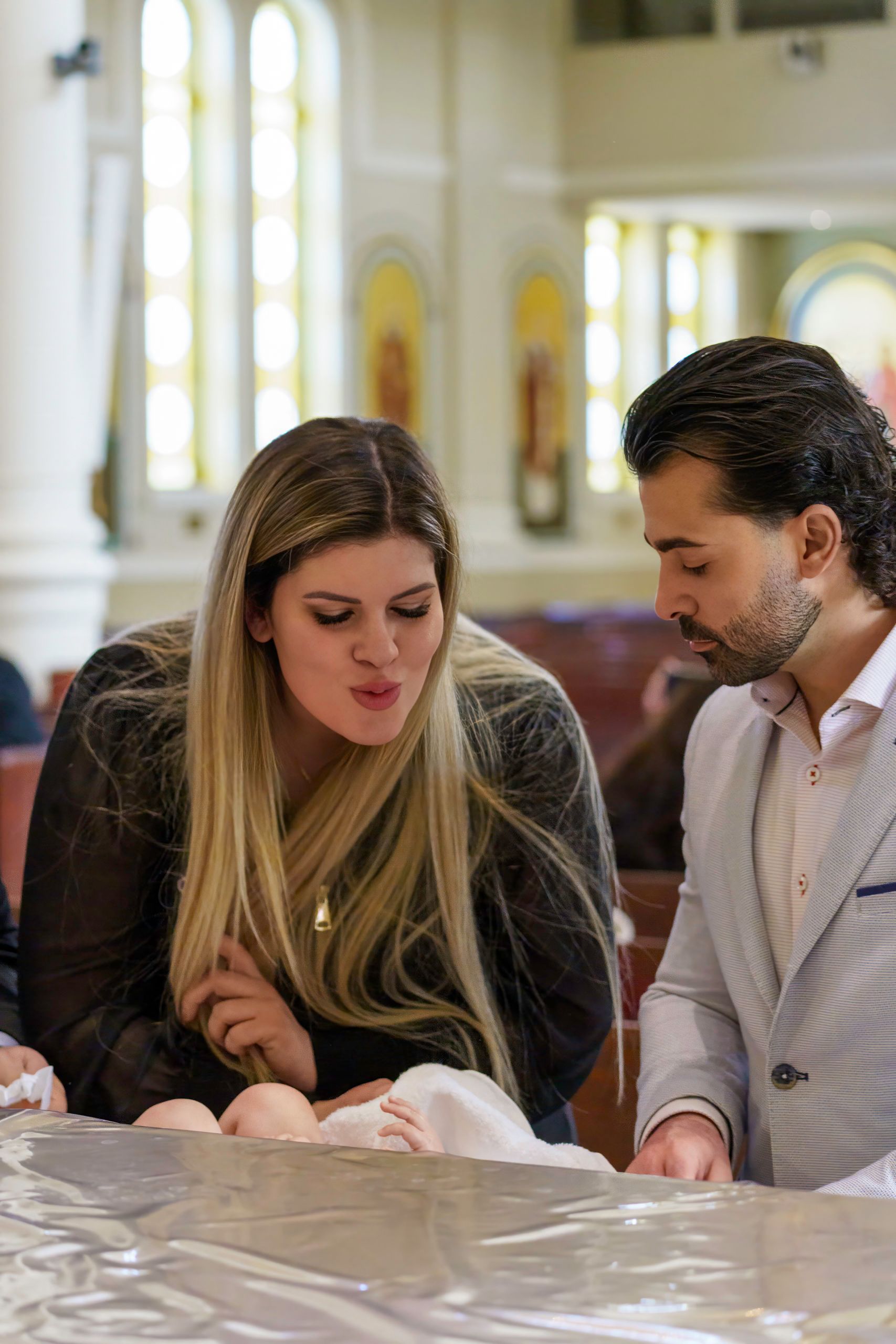 Woman and man lean over a baby in a church, looking down, light streaming in.