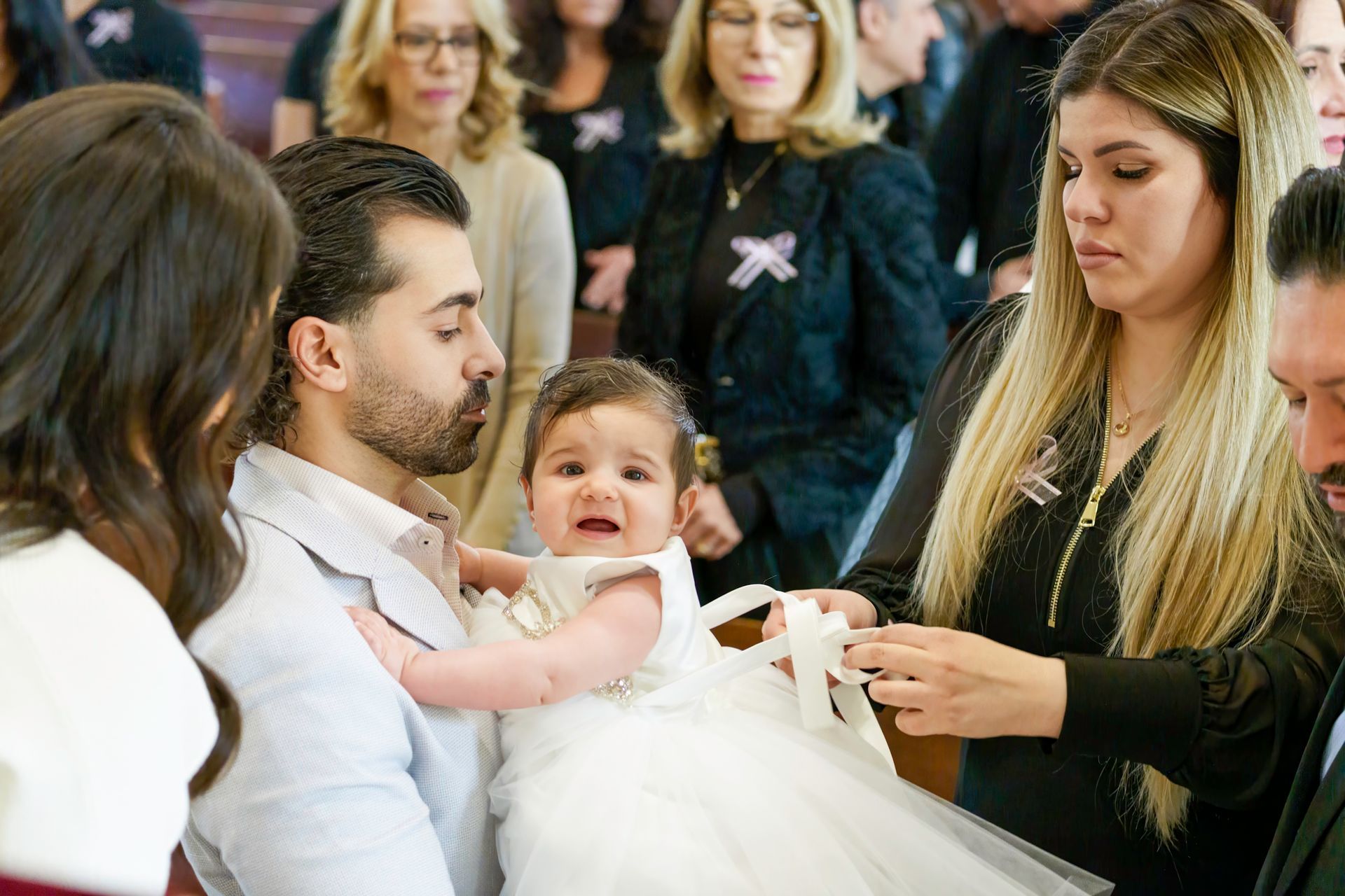 Man holding baby in white dress during ceremony; woman adjusts ribbon. Others observe in background.