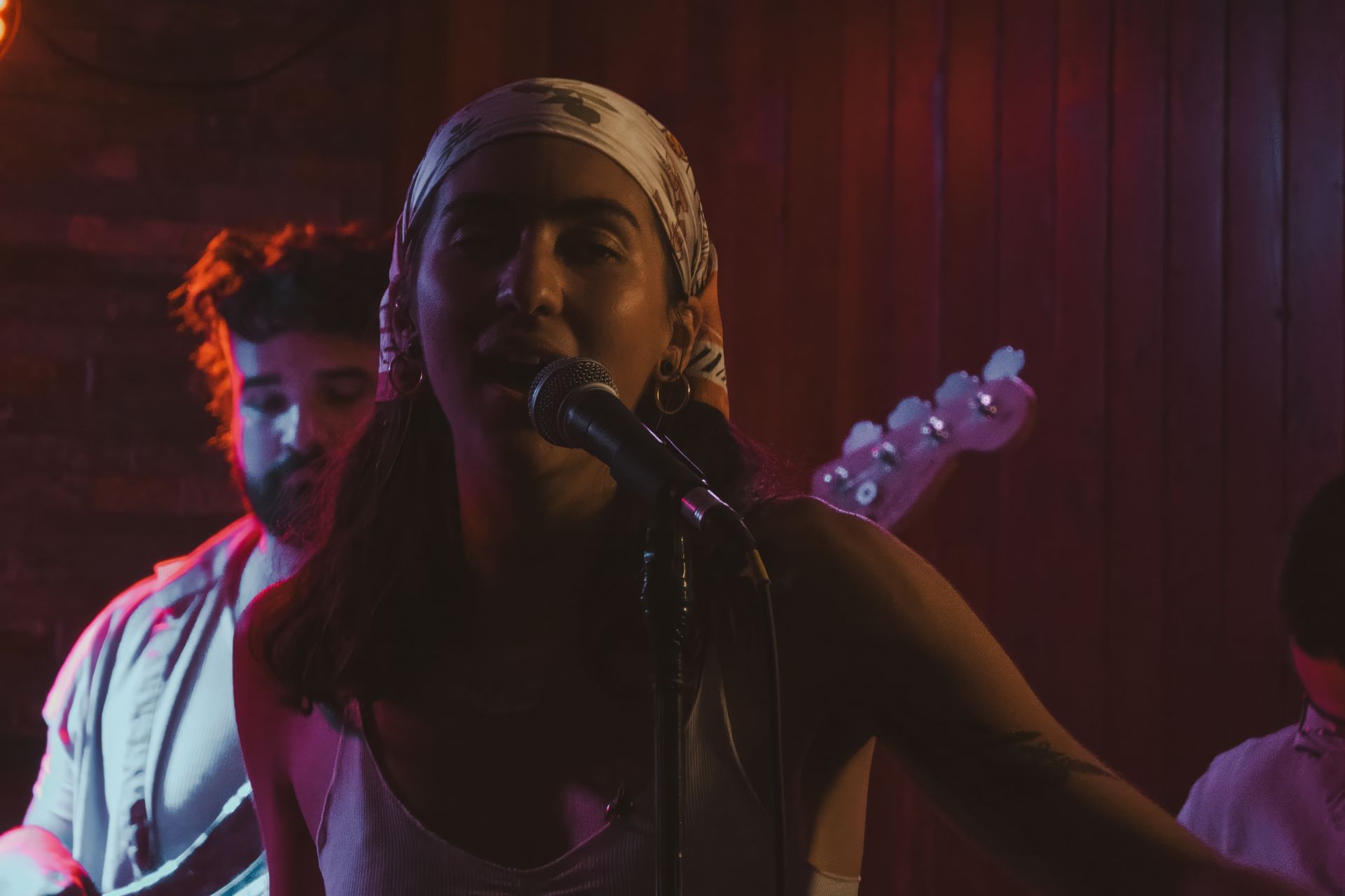 Band performing live on stage, vocalist with headscarf sings into a microphone. Red and purple lighting.
