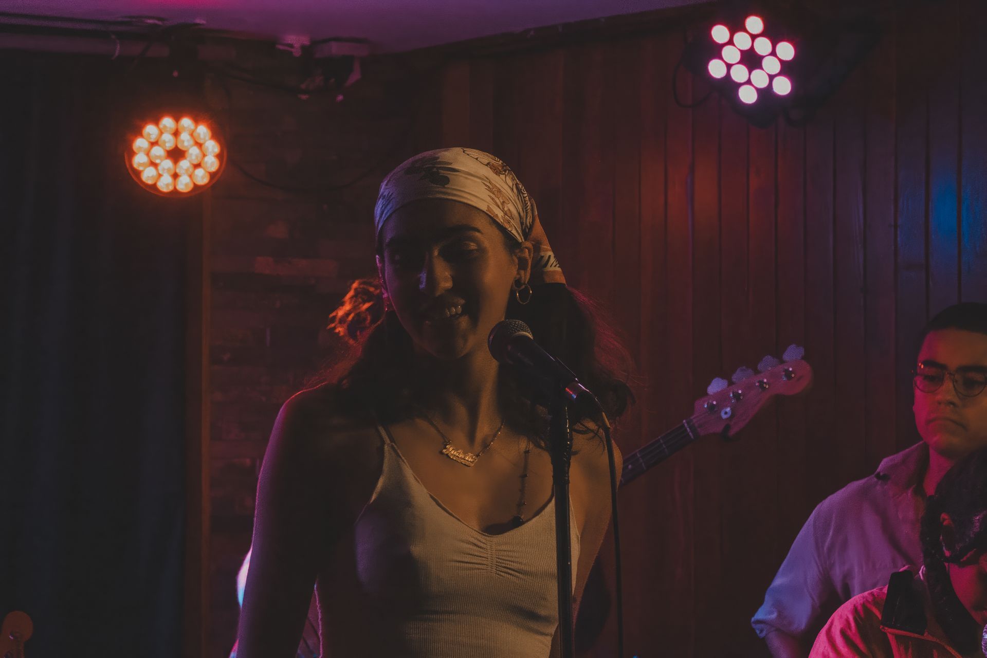 A woman in a white tank top sings into a microphone on a stage with a band, lit by colorful stage lights.