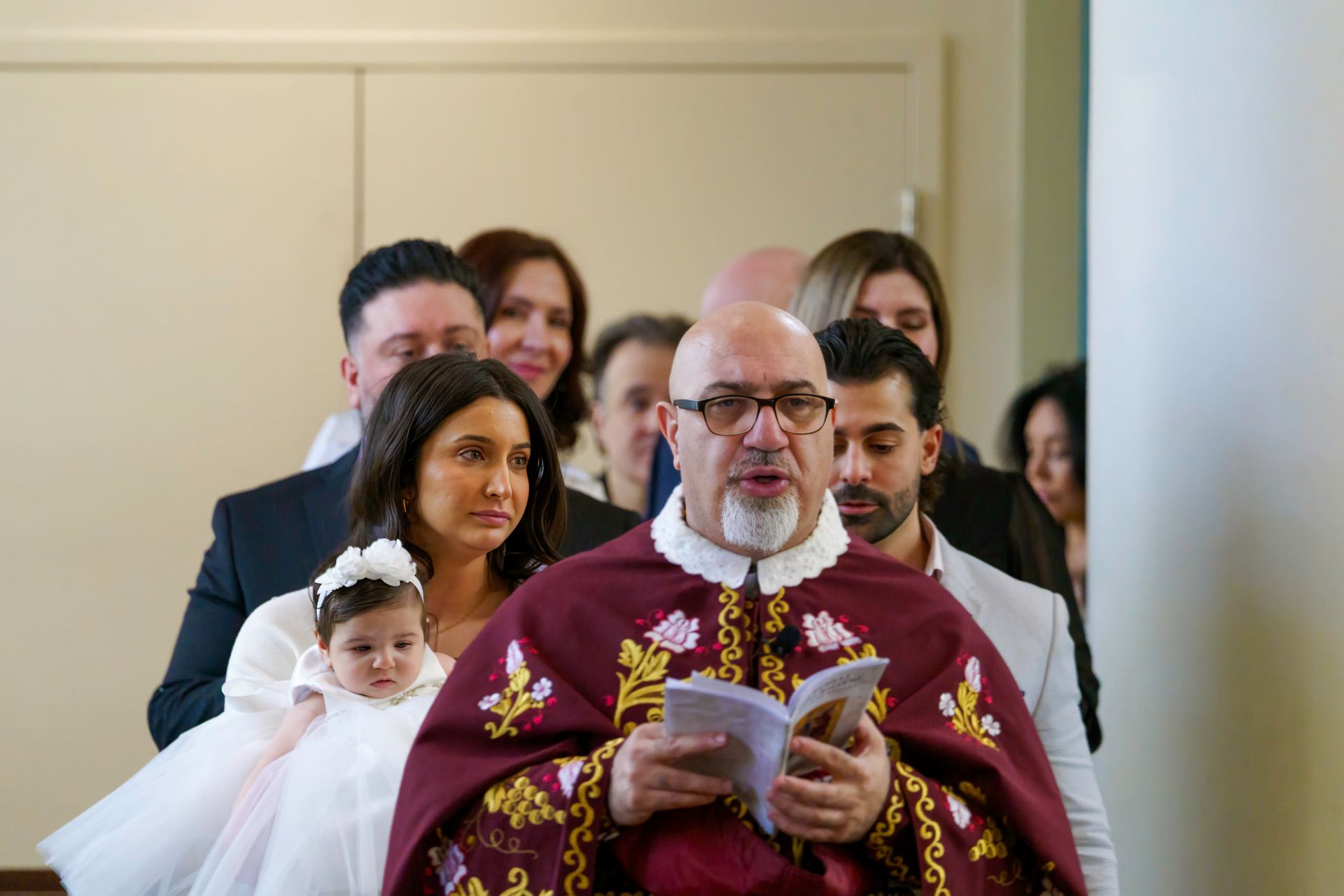 Priest reading from a book during a baptism ceremony; family members in background.
