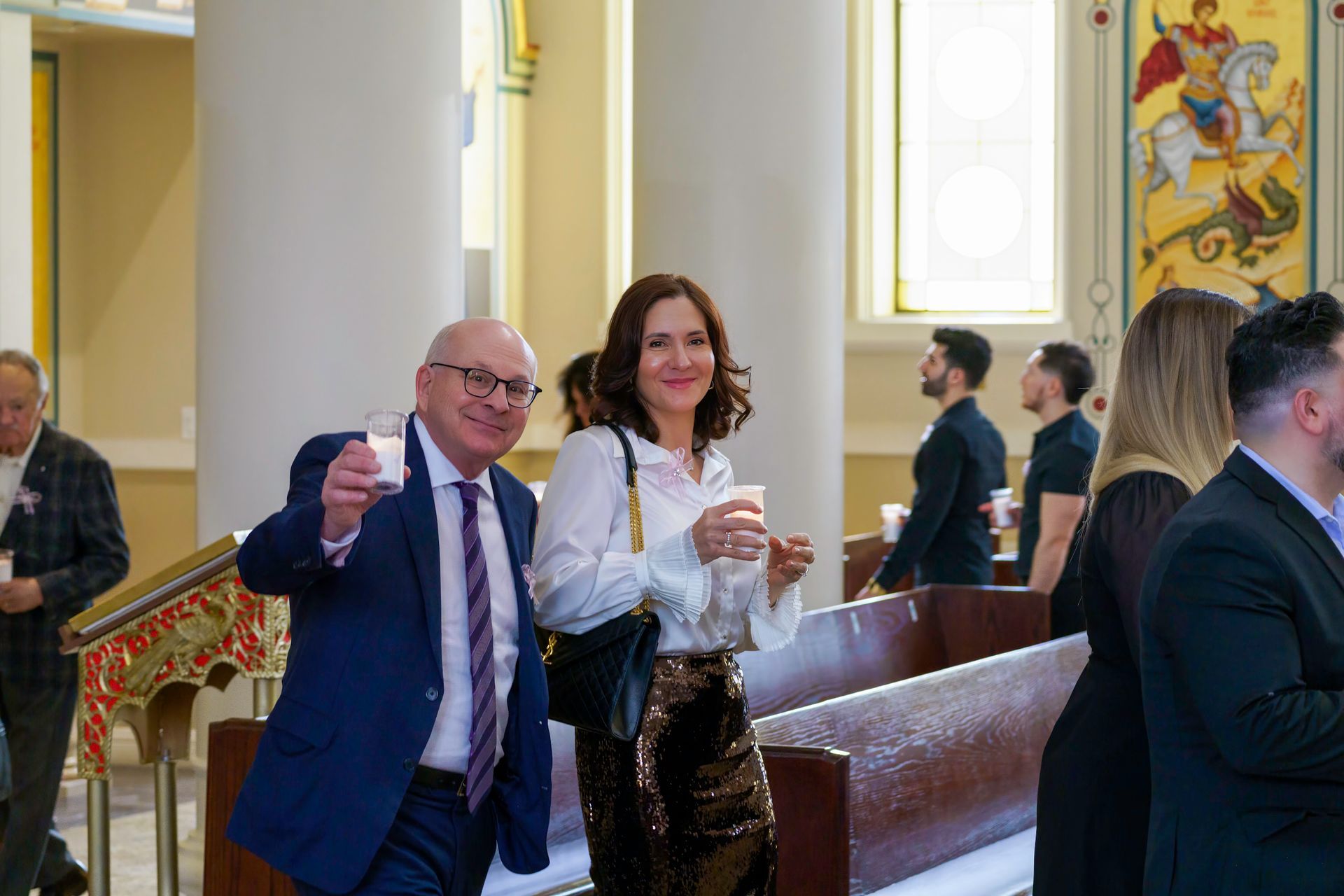 Two people holding candles in a church. Man in a suit gestures with his candle, woman smiles. Other people in the background.