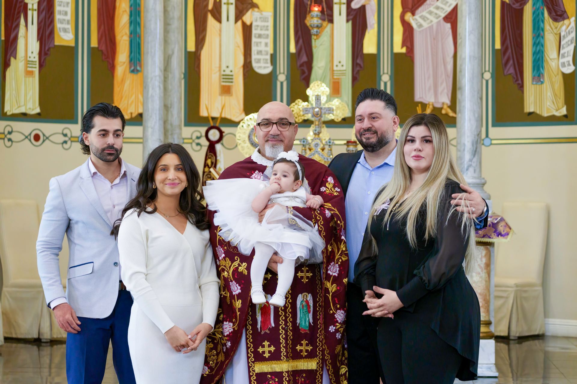 A priest holds a baby, surrounded by family at a baptism ceremony in a church.