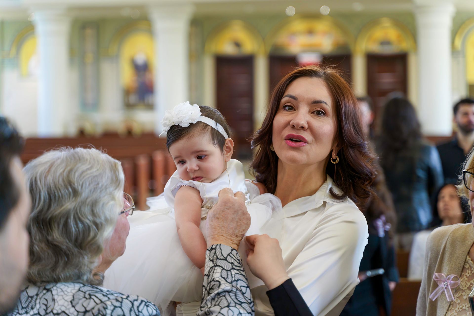 Woman holding a baby in a church; baby in white dress, looking away; other people in background.