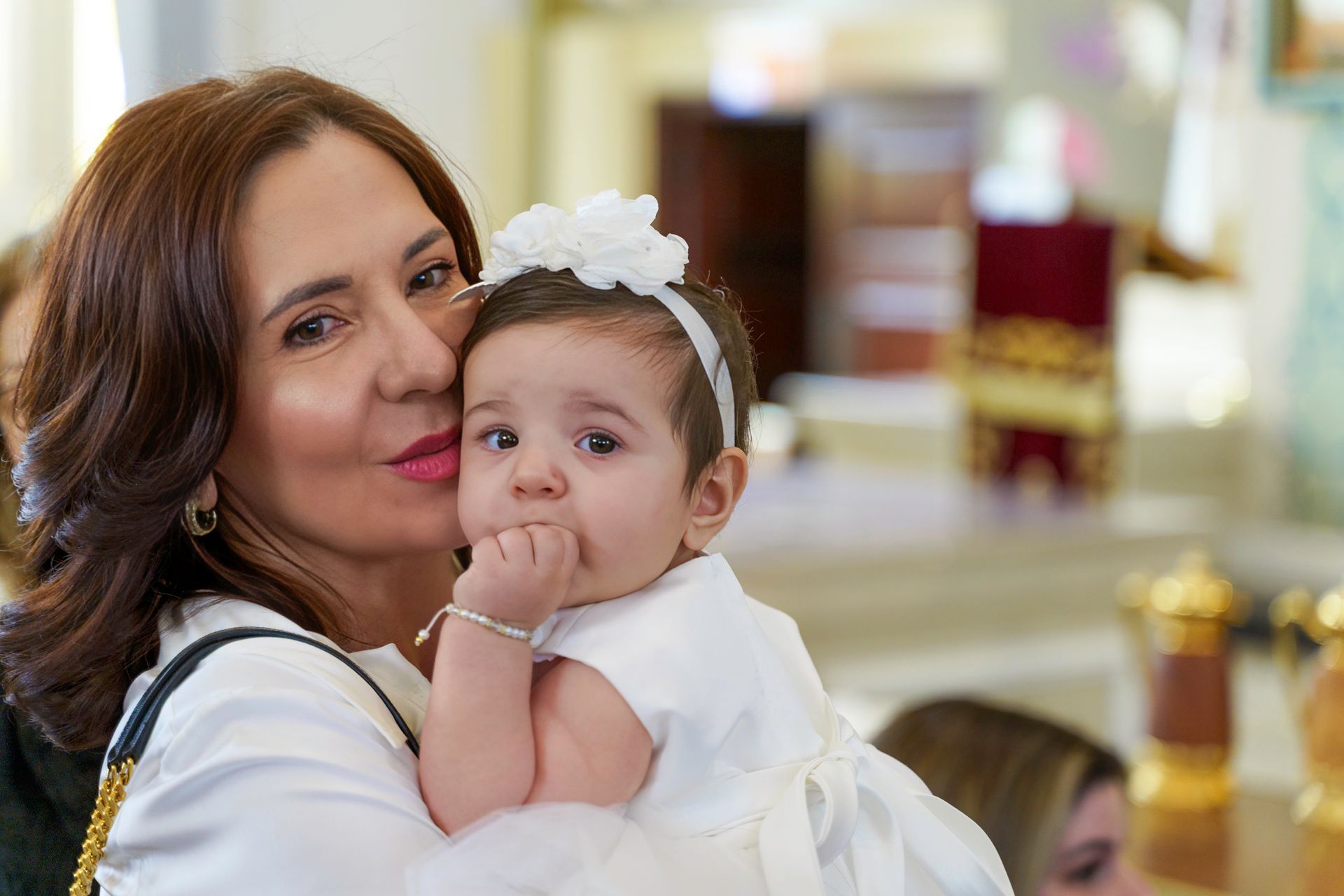 Woman holding a baby, both looking at the camera. The baby wears a white dress and headband, and the woman kisses the baby.