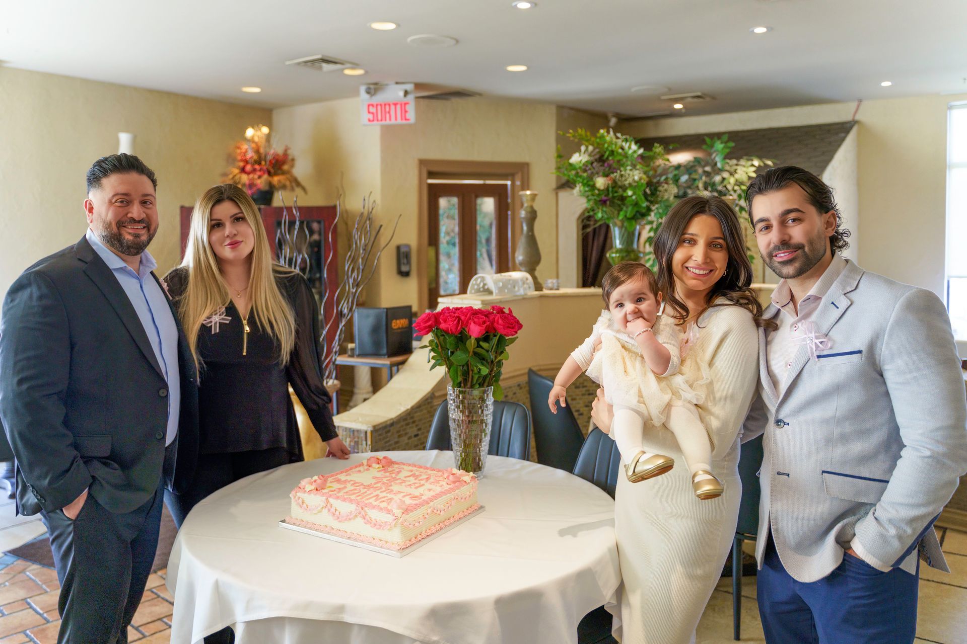 Group of five people around a table with a cake and roses; indoor celebration.