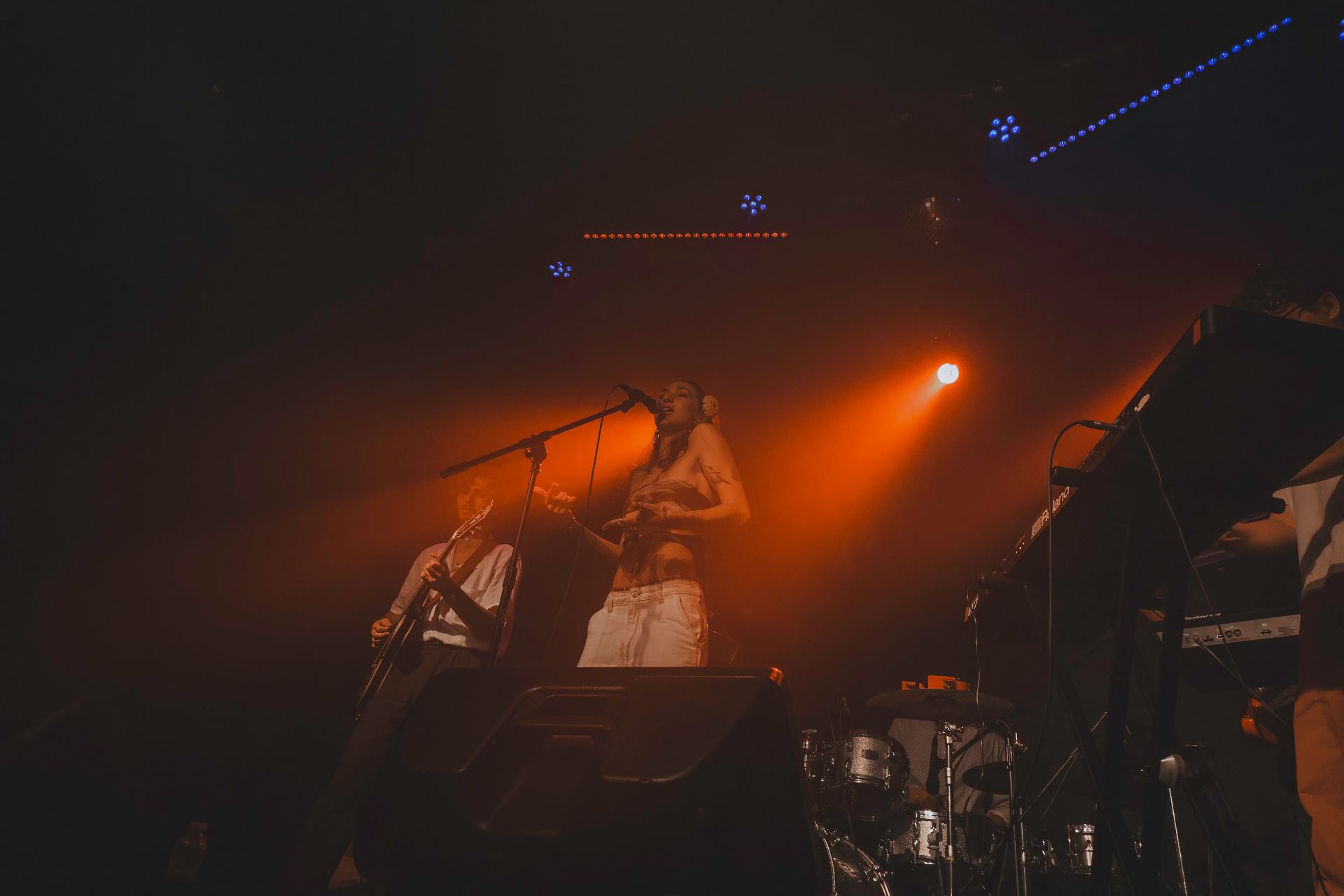 Band performing on stage, bathed in orange light. Musicians play instruments, including a keyboard and drums.