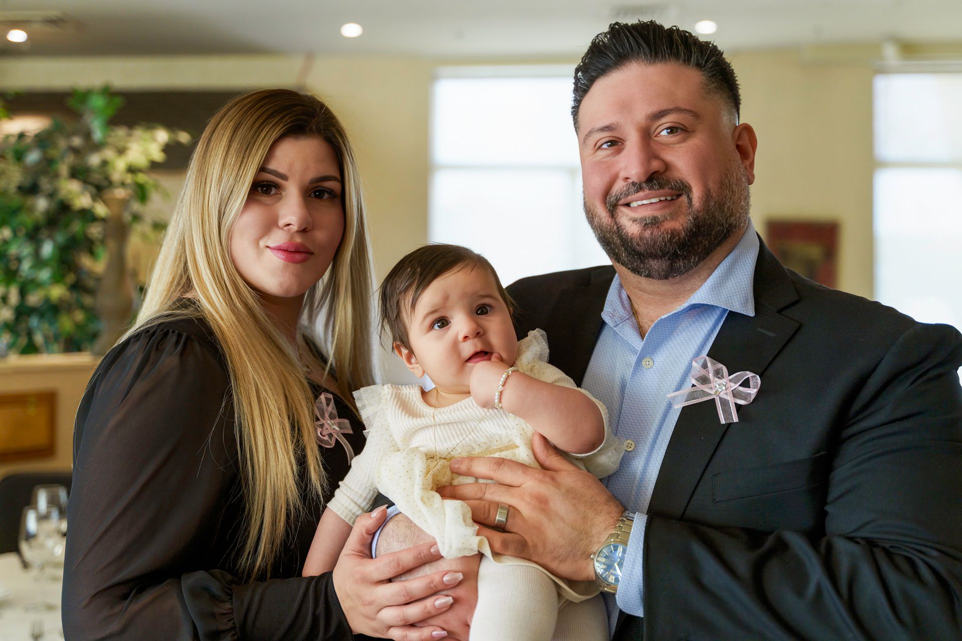 Family of three smiling, posing indoors: Woman, man holding baby in white dress.