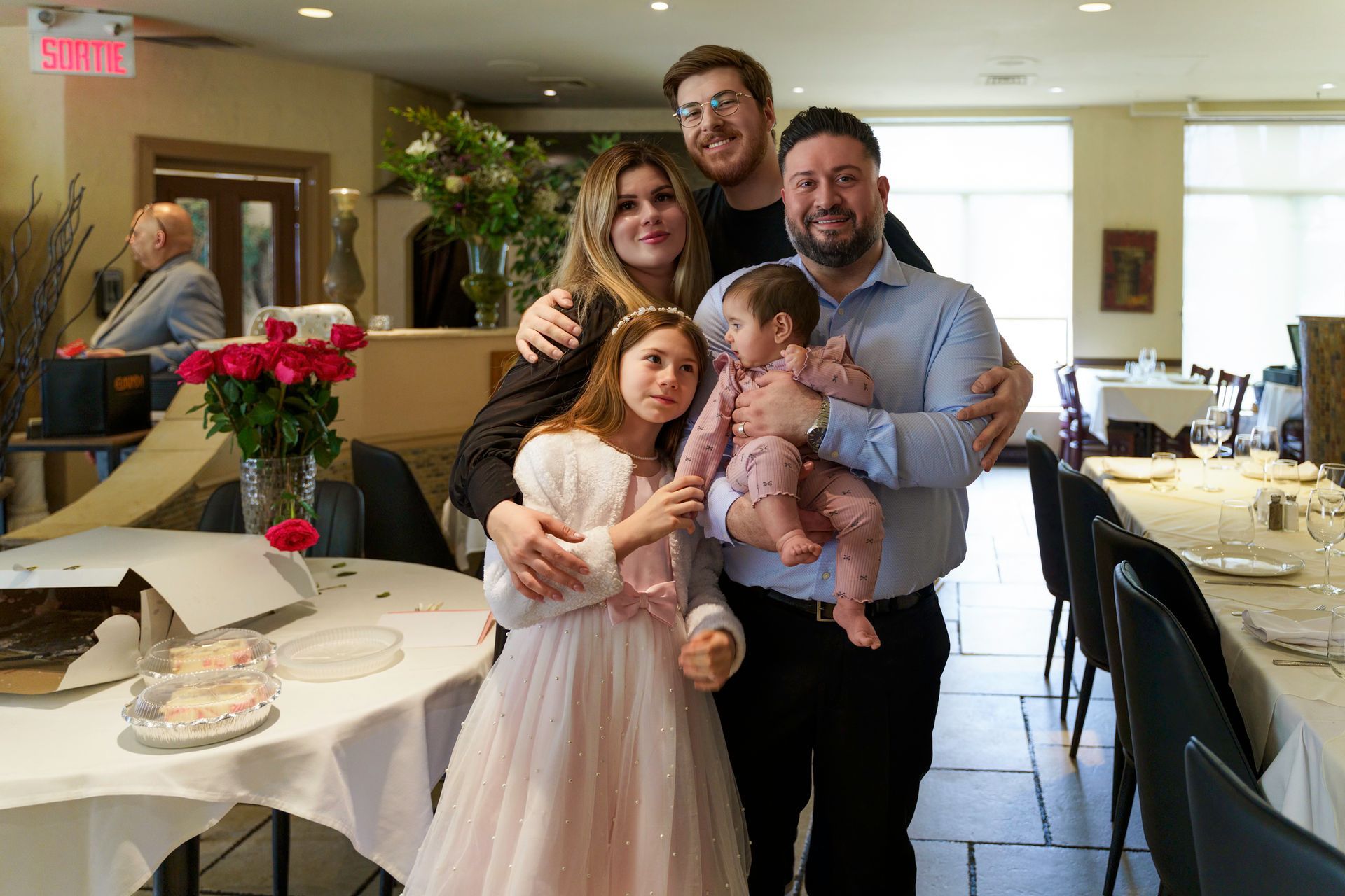 Family of five posing together in a restaurant with a table and roses.