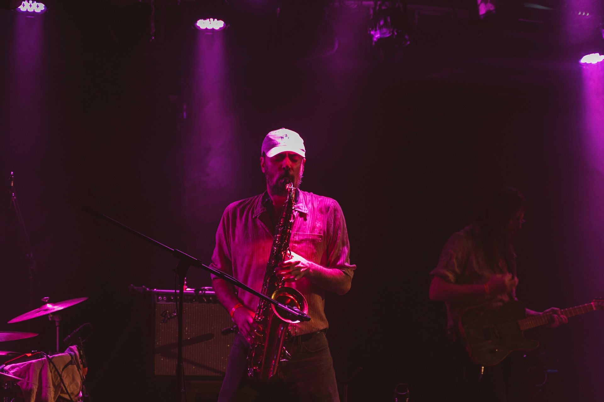 Saxophone player on stage under pink lights. Another person plays guitar in the background.