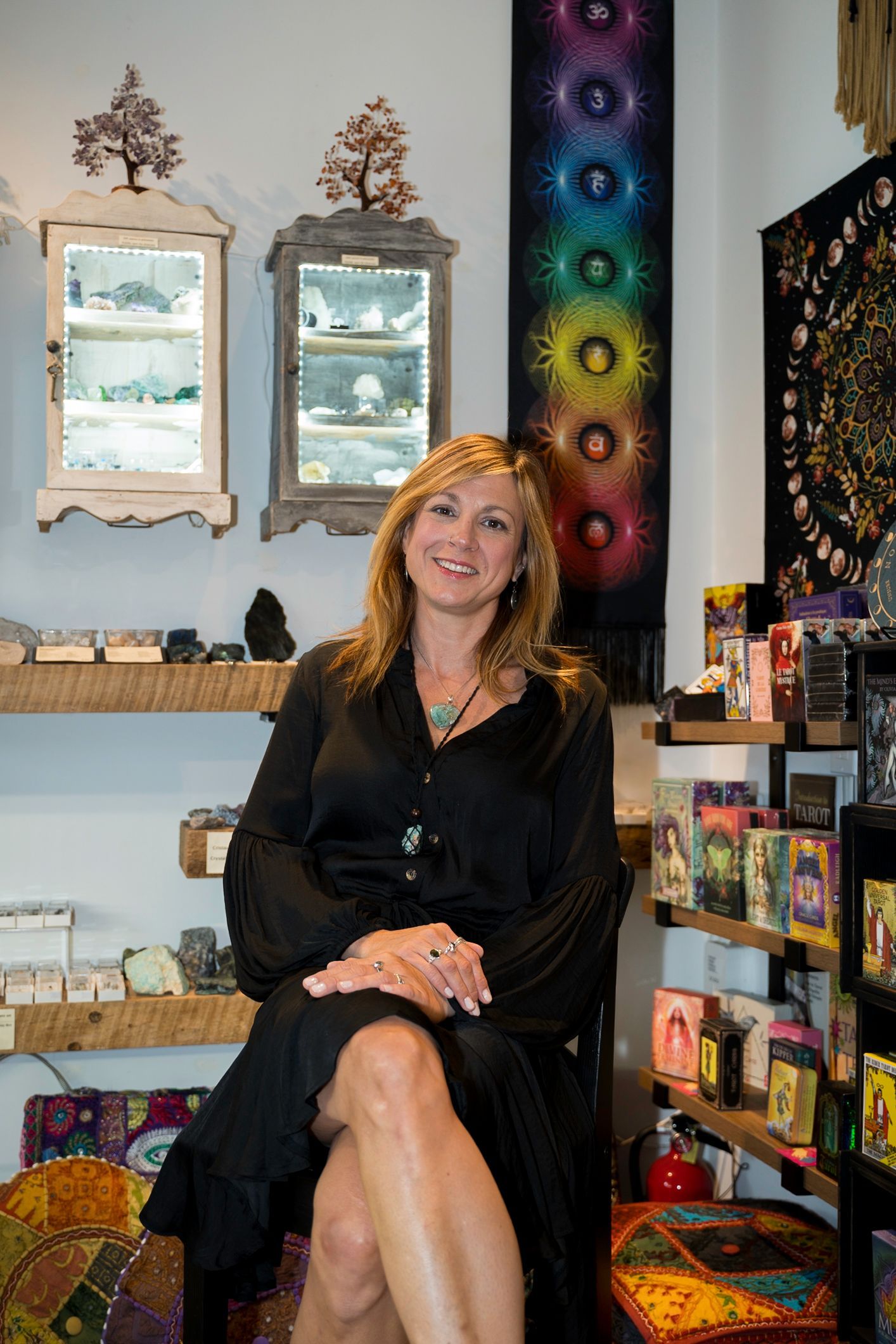 Woman in a shop, seated with legs crossed, in front of shelves with crystals and spiritual items.