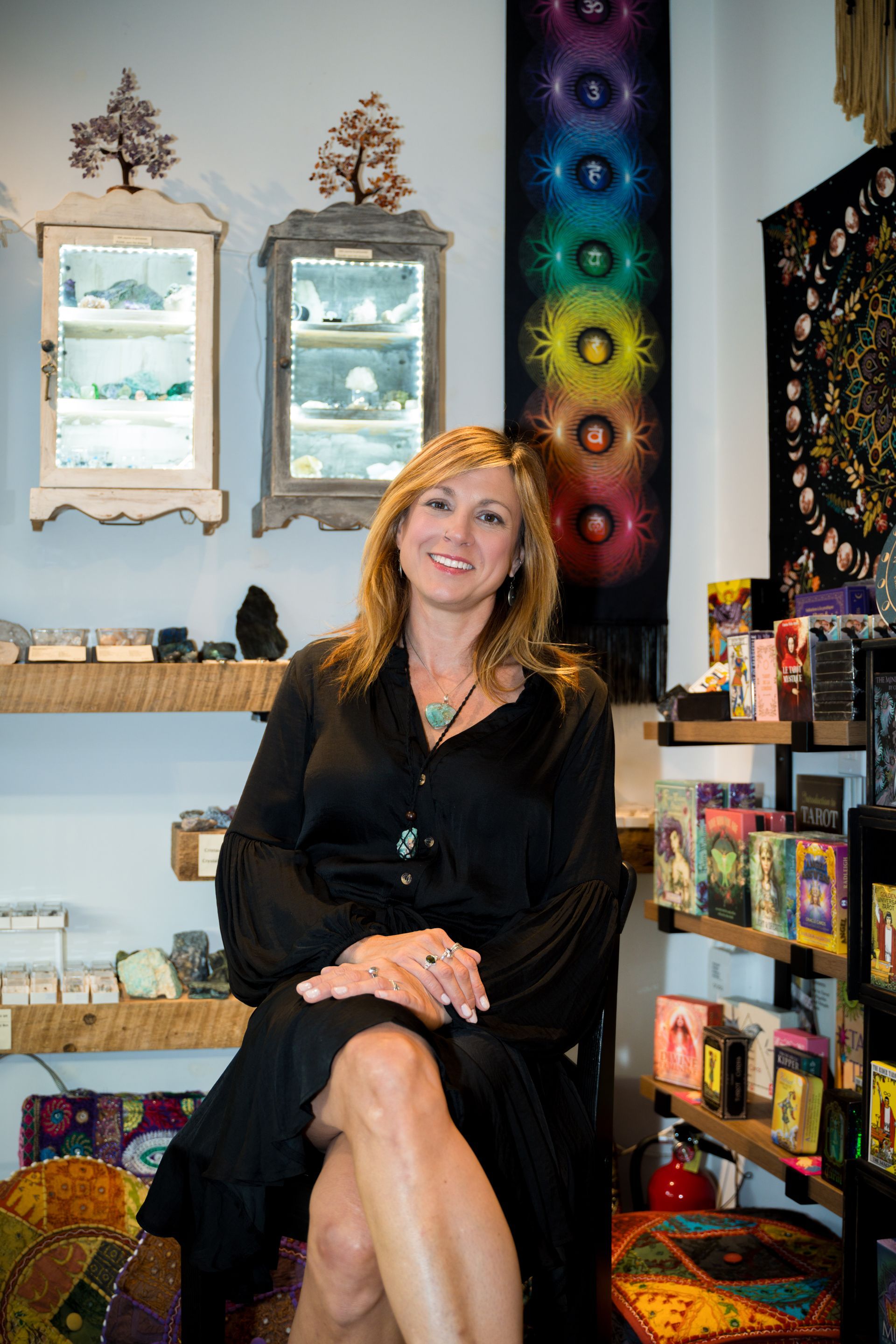 Woman seated in a shop, smiling. Black dress. Shelves with crystals and art. Chakra tapestry in the background.