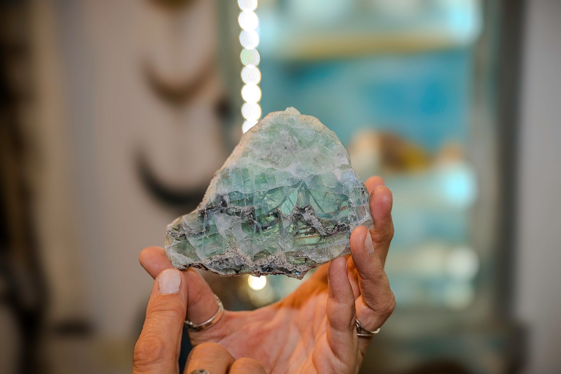 Hand holding a rough, green-hued rock with dark veins, blurred background with white lights.