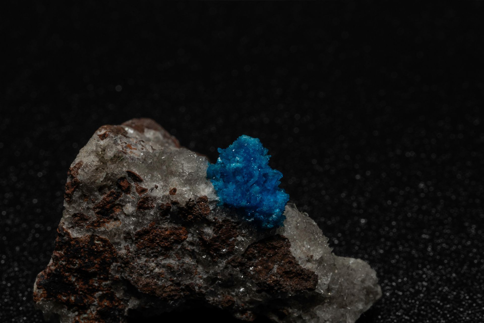 A vibrant blue crystal cluster sits on a small rock with brown and gray sections against a dark background.
