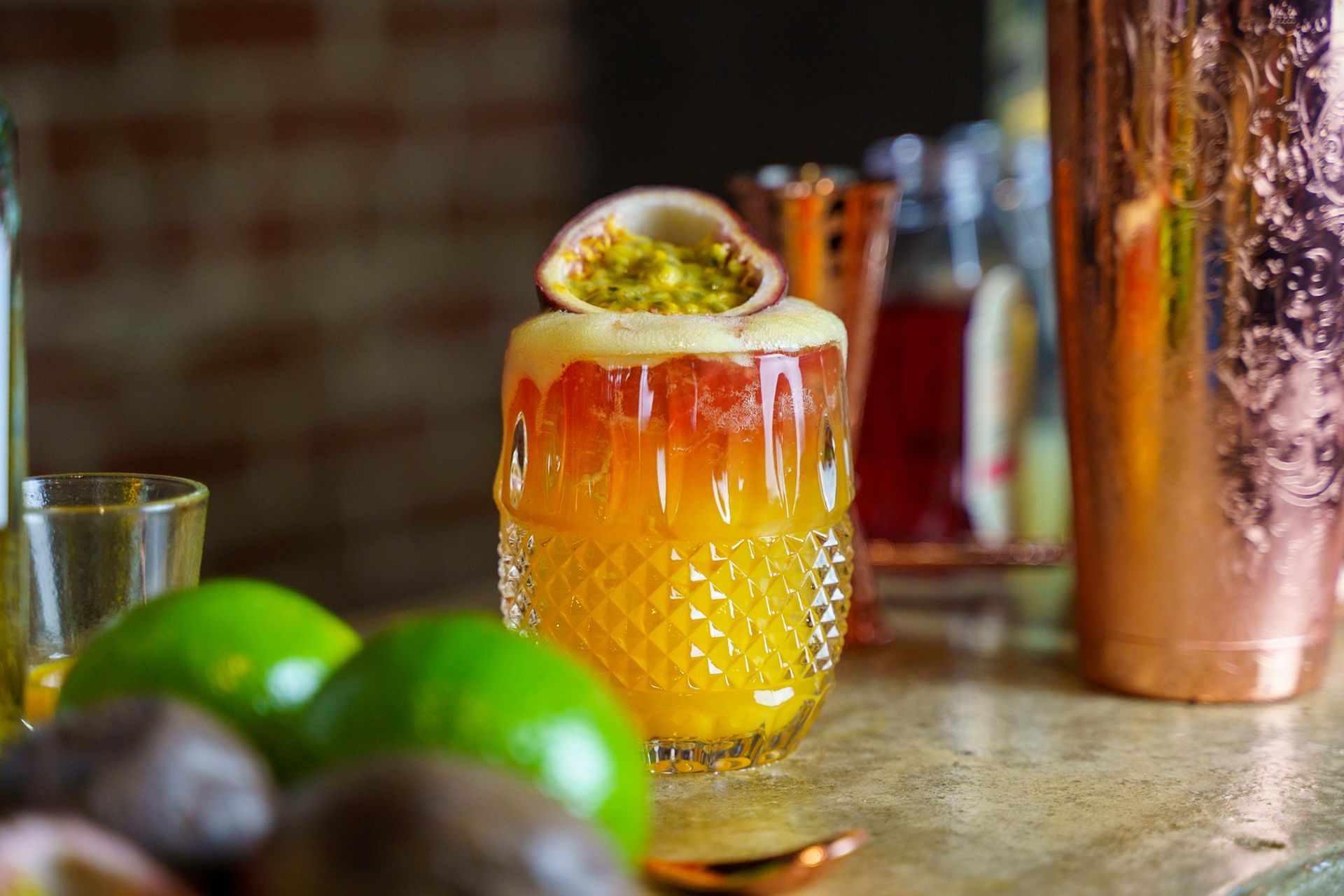 Cocktail in a cut-glass with a passion fruit garnish, next to limes and a copper shaker.
