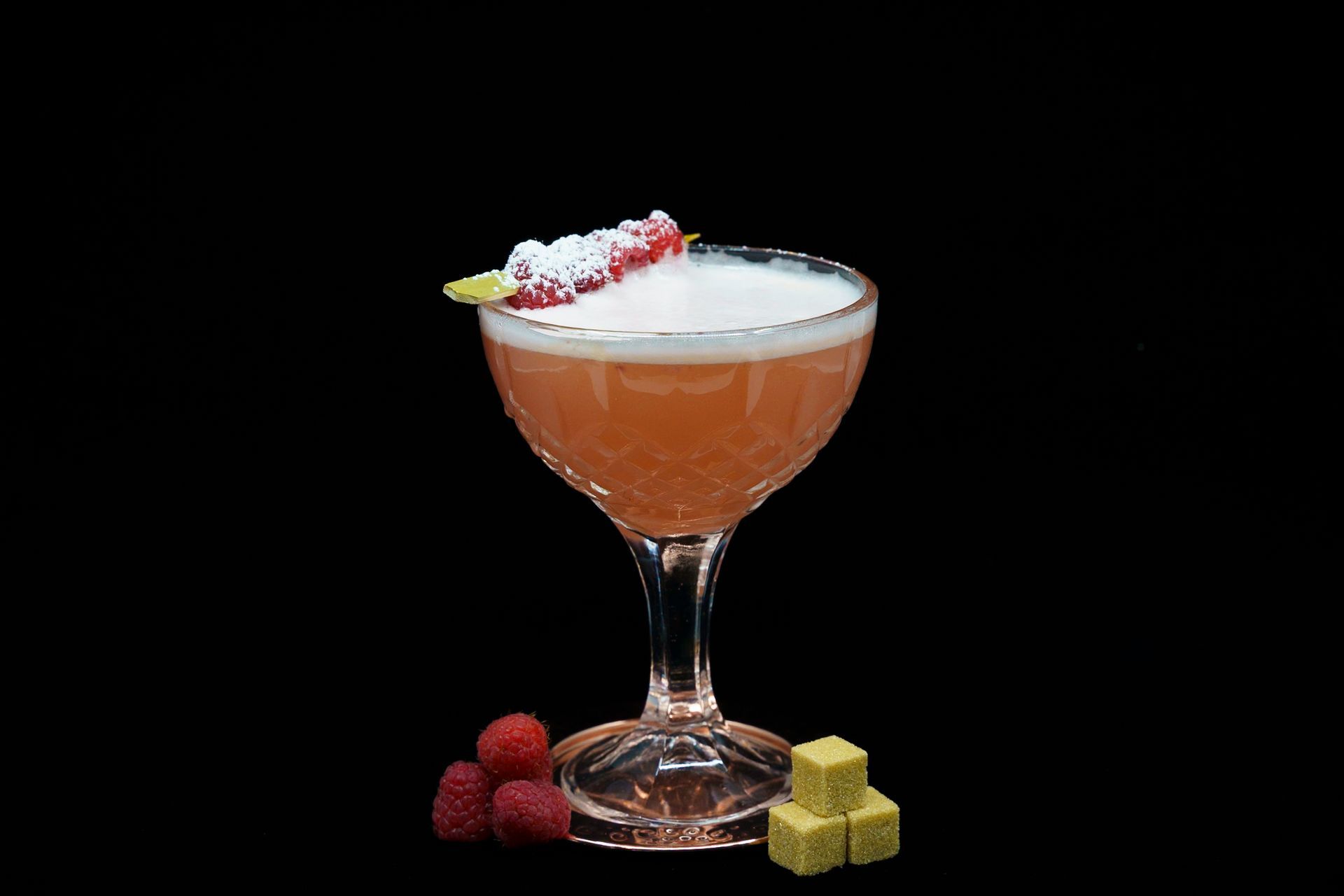 Pink cocktail in a stemmed glass, garnished with raspberries and a lime wedge, on a black background.