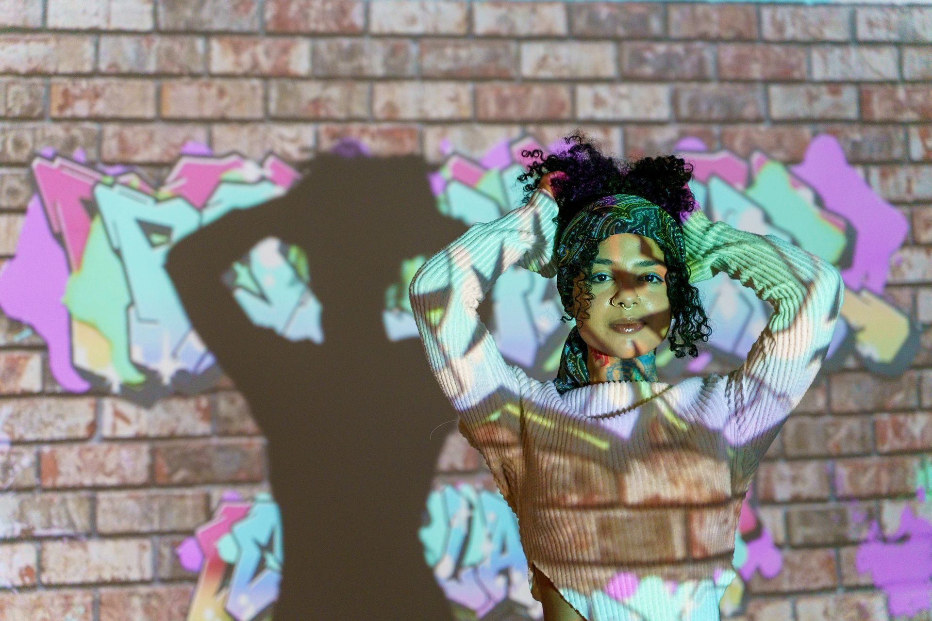 Woman with arms raised, lit by graffiti projection on a brick wall.