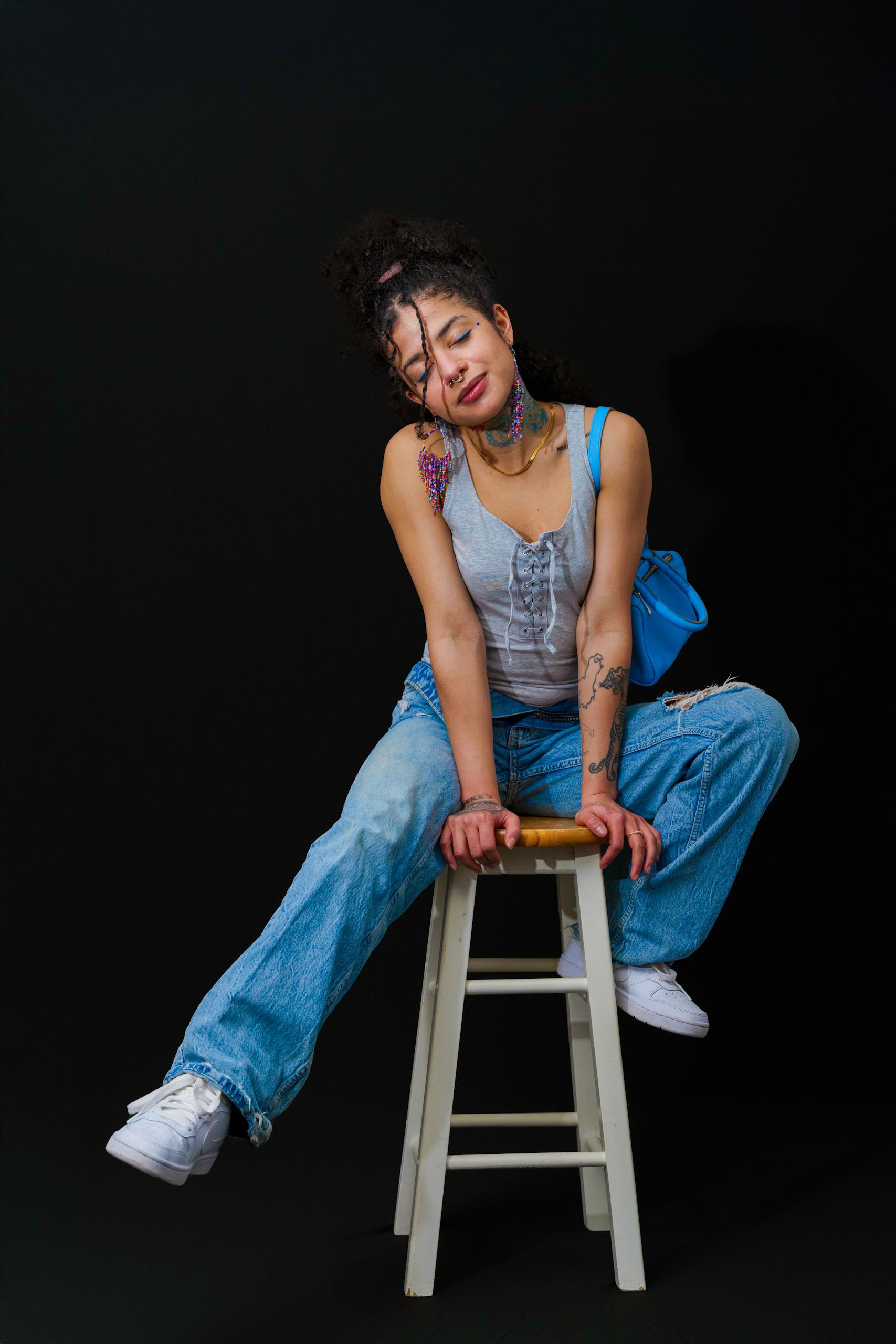 Woman in denim outfit, posing on stool against black background, smiling.
