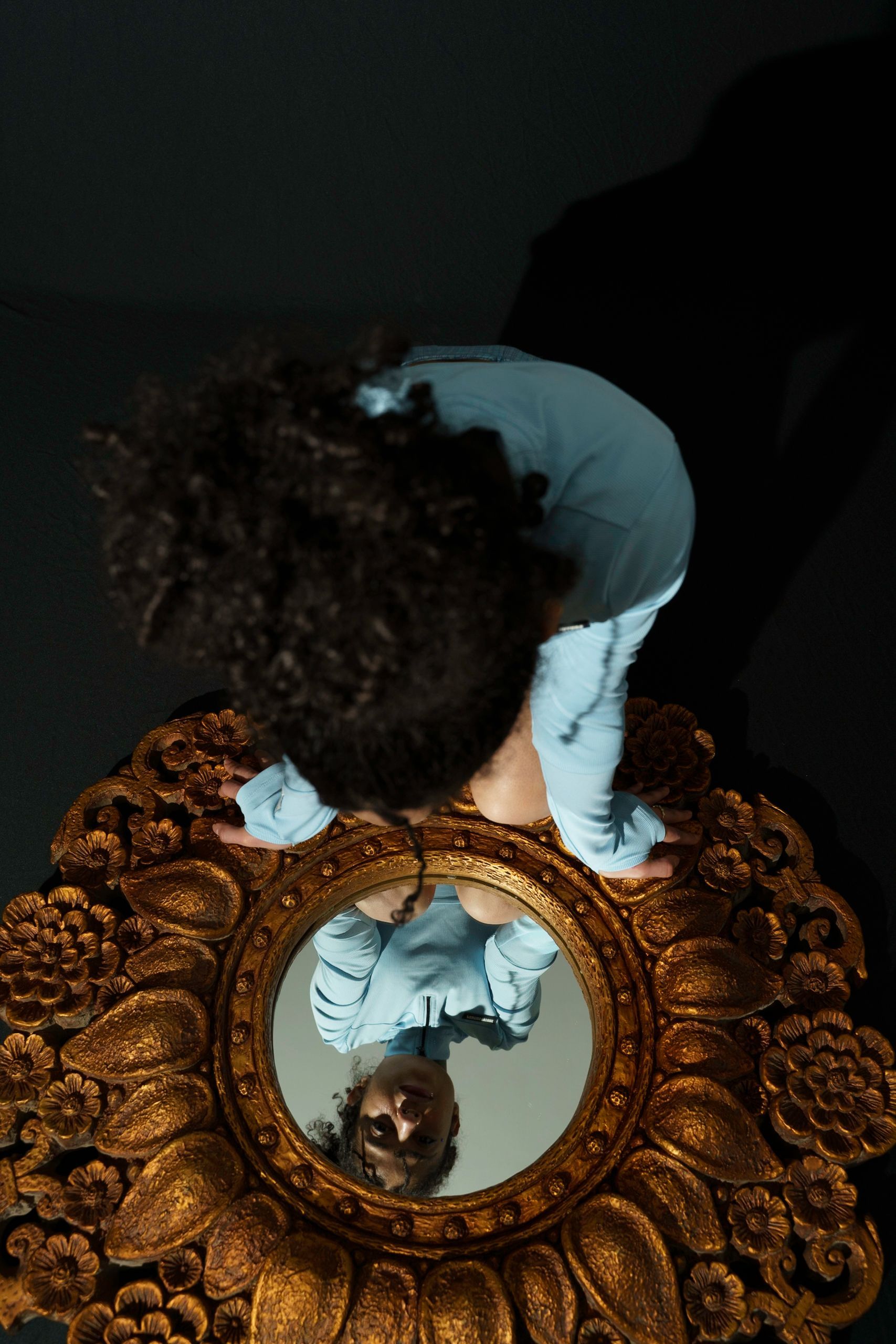 Person leaning over ornate gold mirror, looking at reflection. Dark setting, wearing blue.