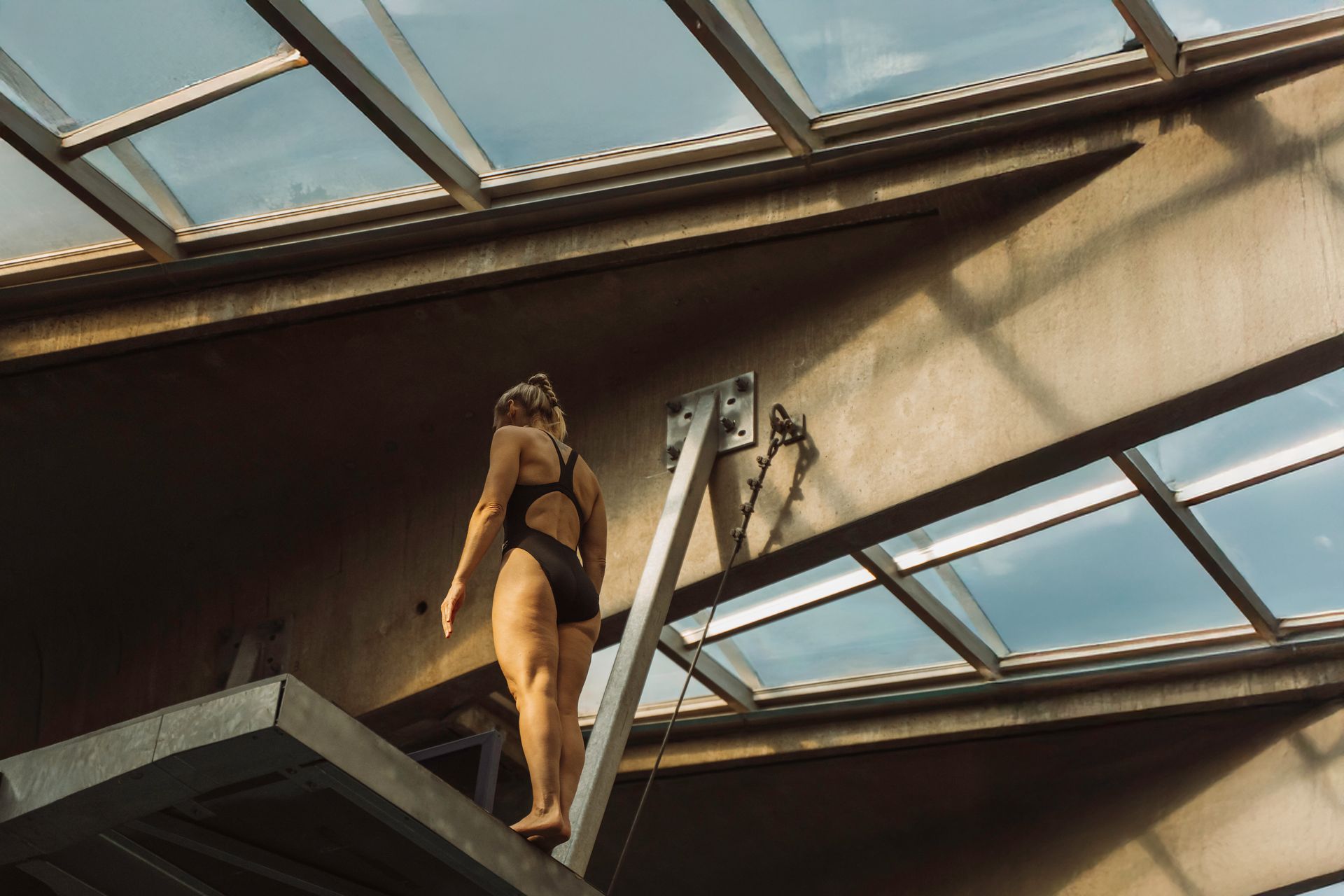 Woman in black swimsuit stands on a diving board, preparing to jump into a pool. Indoors, natural light.