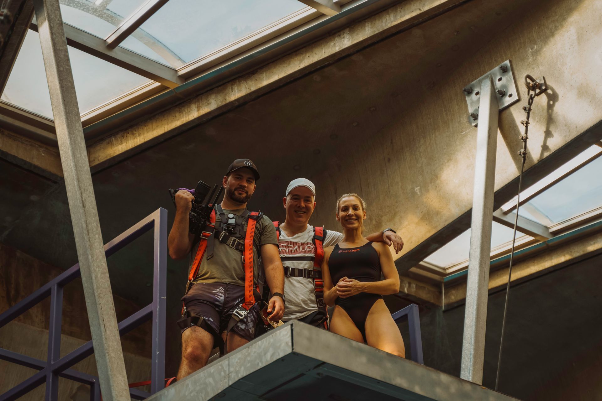 Three people pose on a platform beneath a skylight. Two men wear harnesses, one in a cap, the woman in a swimsuit.