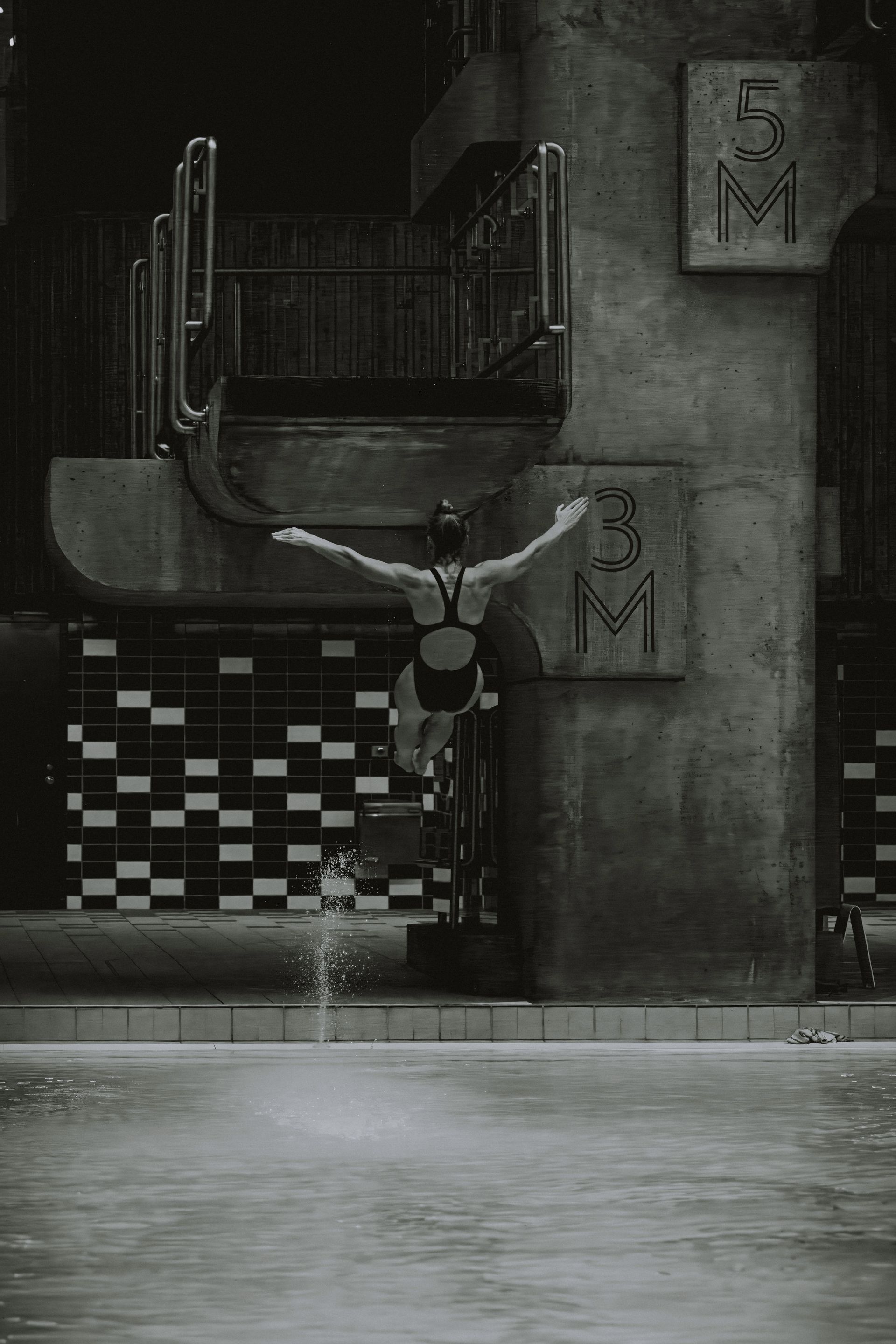 Diver in mid-air above pool. 5M sign on concrete diving platform. Black and white.