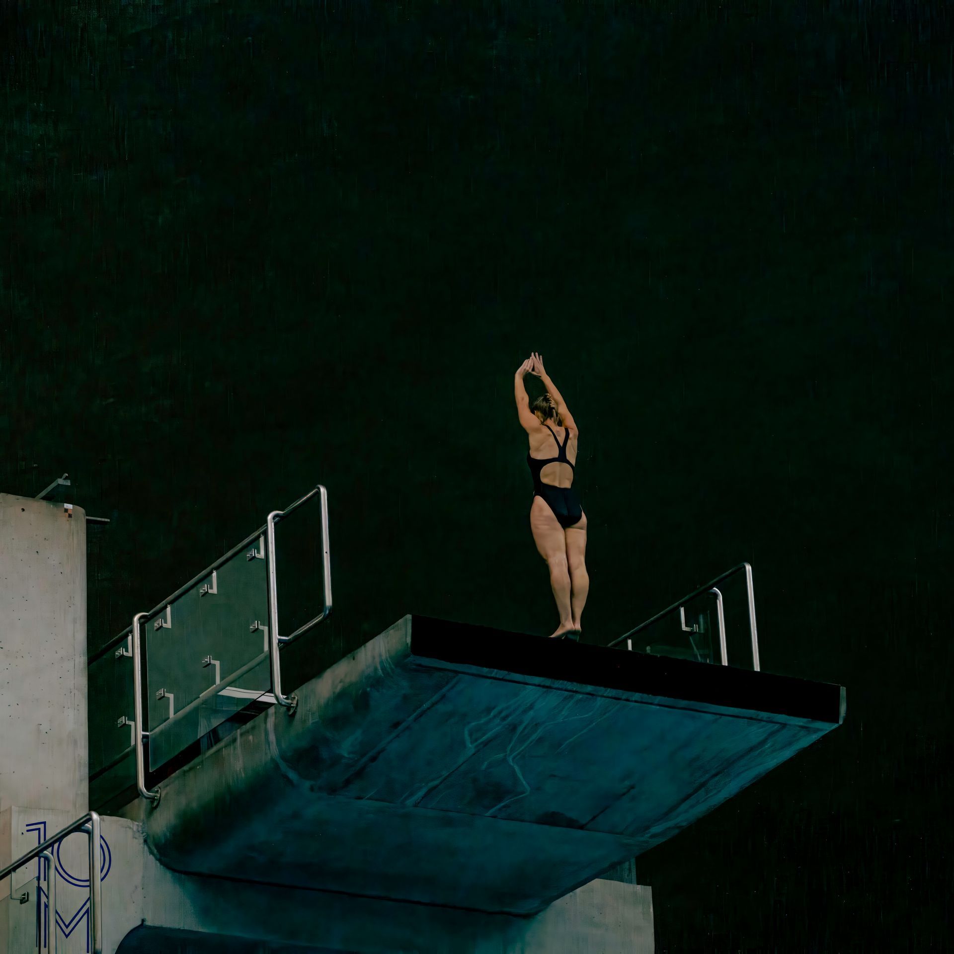 A swimmer stands on a diving board, arms raised, poised to jump into dark water.
