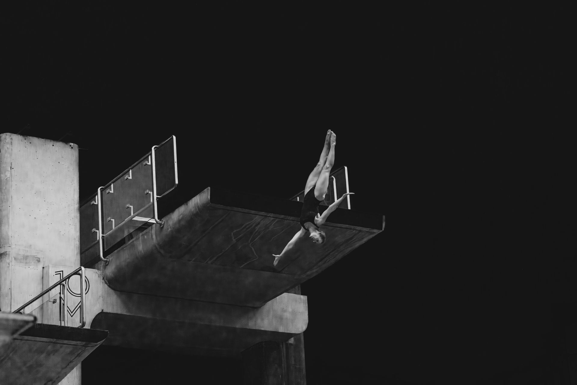 Diver in mid-air above a diving board, silhouetted against a black background.