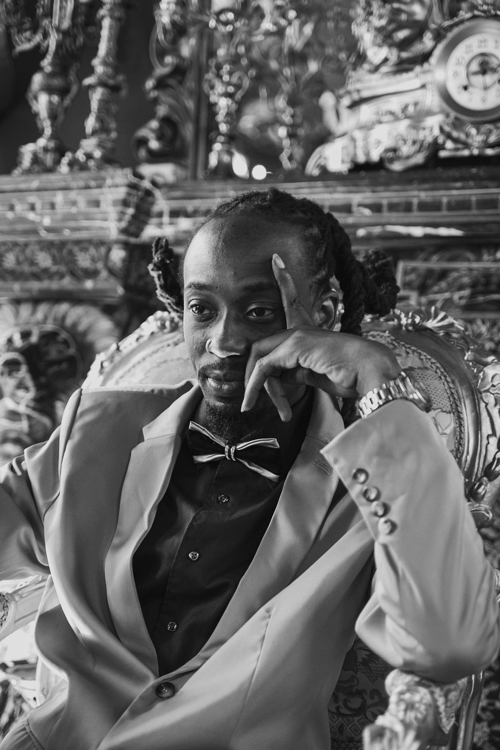 Person in a light suit, resting chin on hand, seated in an ornate chair. Bow tie, clock, and detailed backdrop.