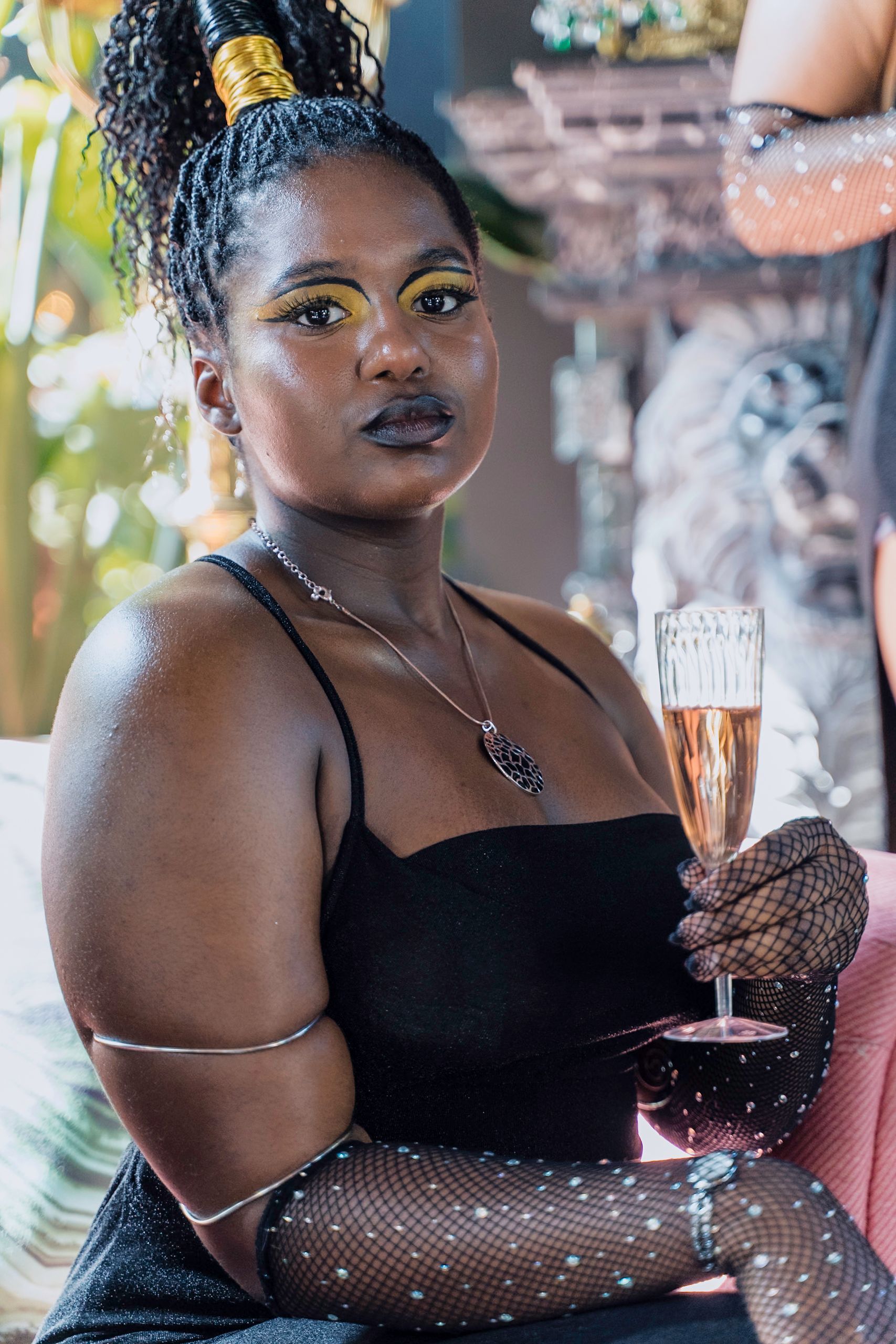 Woman in black dress and gloves holds champagne, gold eye makeup, curly hair in a high bun.