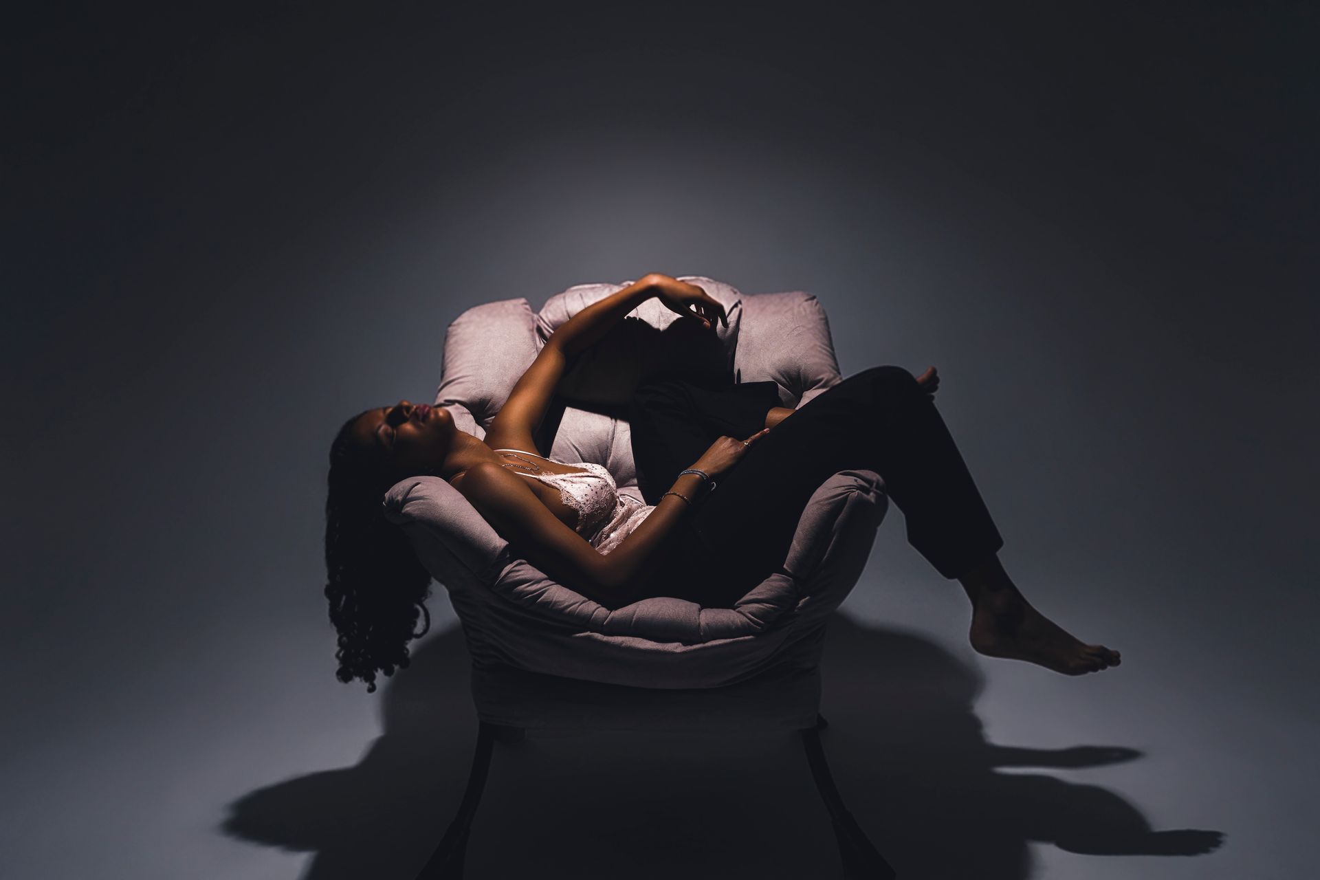 A person lounging in a plush armchair under a spotlight against a dark, minimalist background.