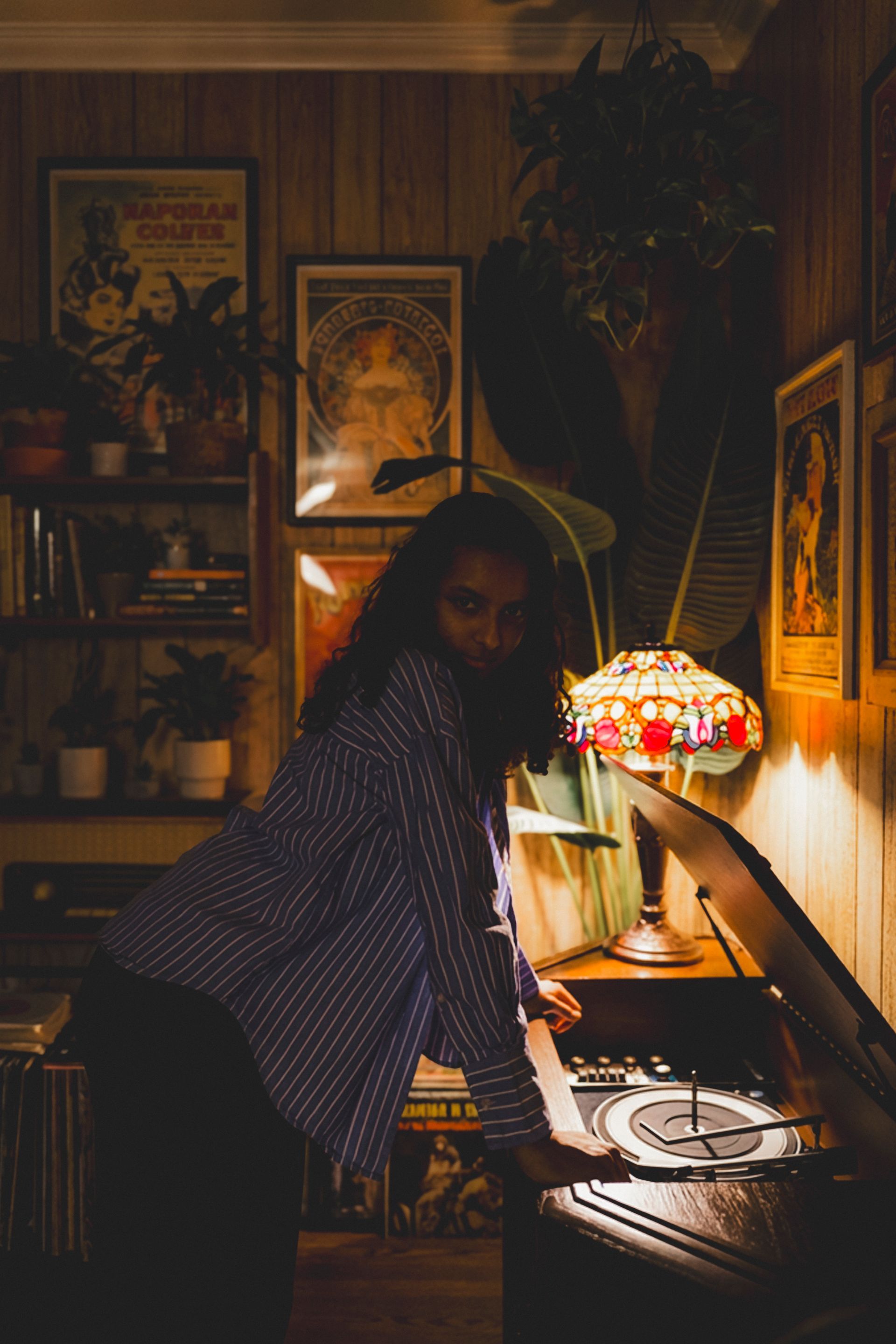 A person in a plaid shirt leans over a record player in a cozy, dimly lit room filled with plants and framed art.
